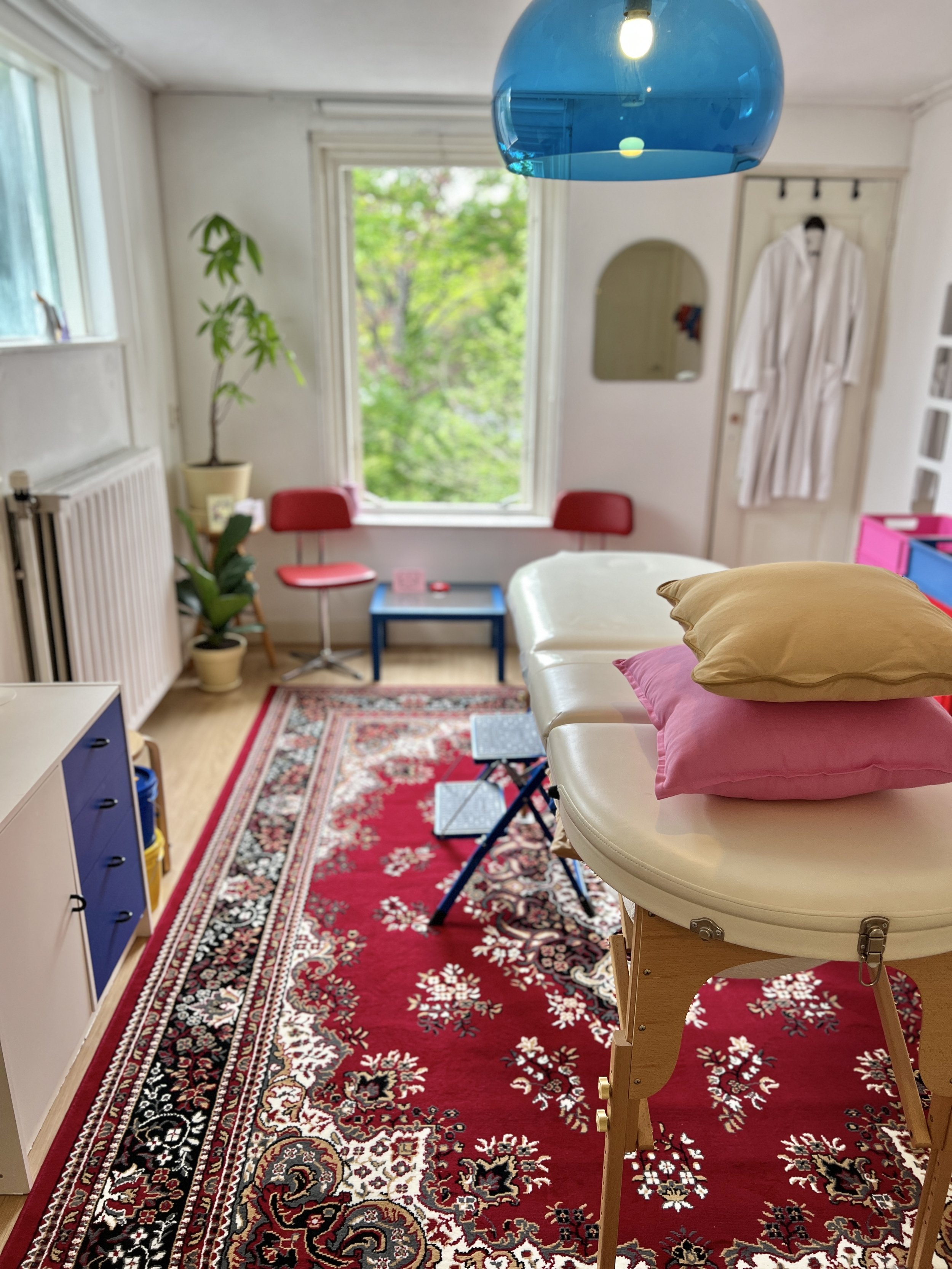 Sfeervol en warm body casting atelier van Venus in Vorm met behandeltafel, twee kussens, stoelen, hanglamp, raam, rood Perzisch vloerkleed, planten en ochtendjas op de achtergrond.