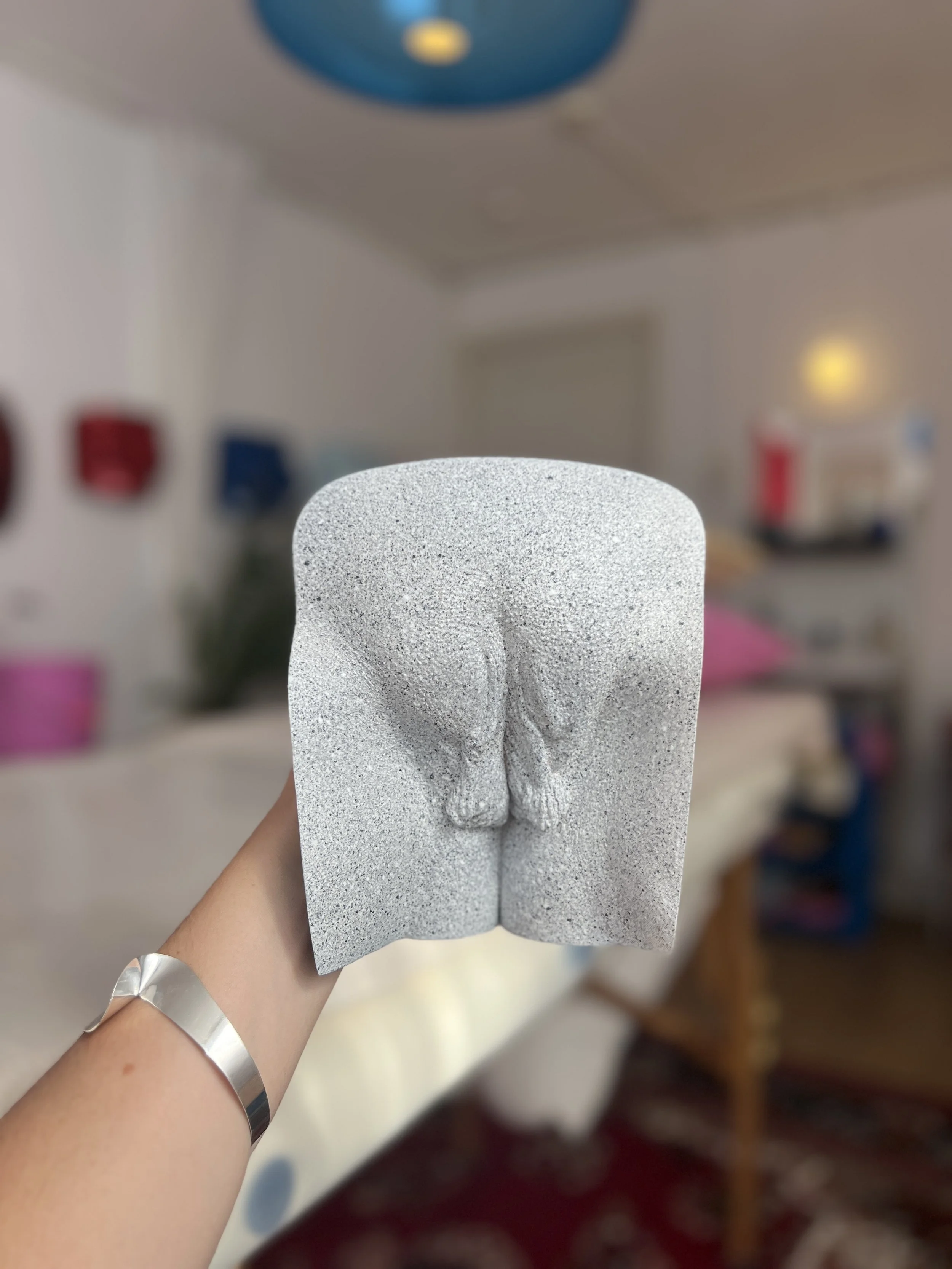 Lichtgrijs vulva-beeld met steeneffect afwerking, handgemaakt van hars, kunstwerk van Venus in Vorm, vastgehouden in het atelier, met zicht op andere sculpturen en behandeltafel op de achtergrond.