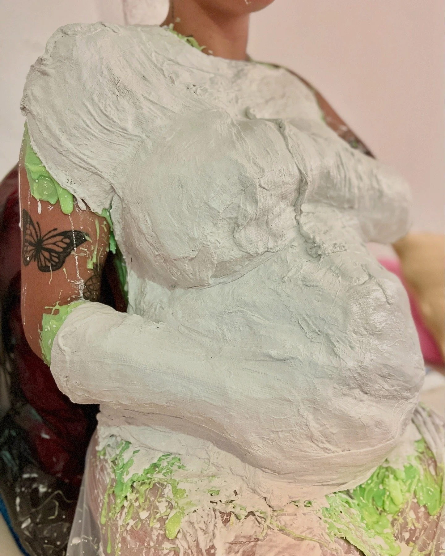 Vrouw tijdens zwangerschap body casting bij Venus in Vorm met groene siliconen over haar buik, borsten en armen en daarover een steunkap van gips. Draagt schort voor bescherming van ondergoed, zodat het schoon blijft, vlindertattoo zichtbaar op arm.