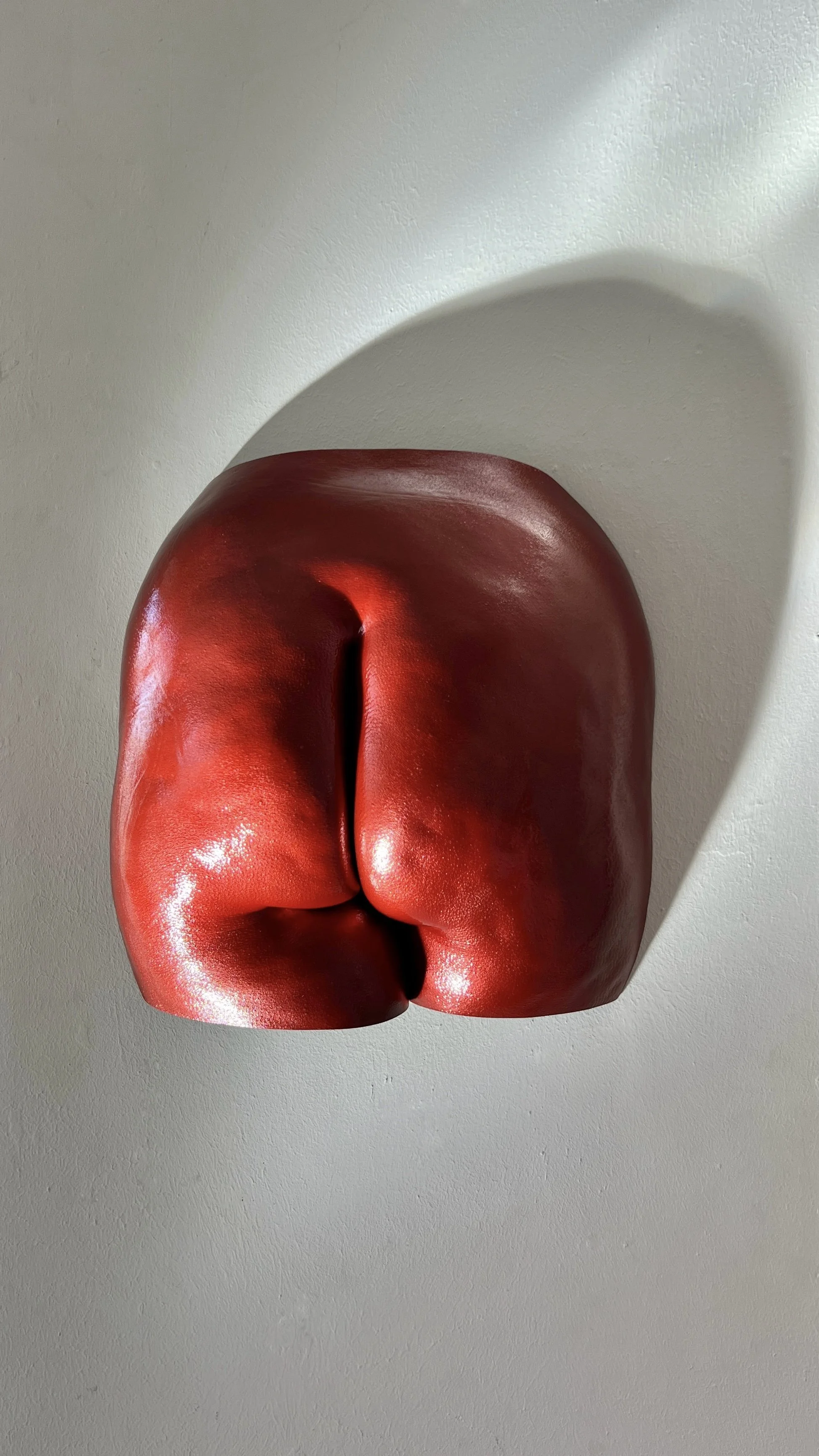 Rood metallic beeld van billen met zichtbare cellulitis en huidstructuur, handgemaakt van hars, kunstwerk van Venus in Vorm, gefotografeerd in het bodycasting atelier met spotlight die schaduw op de muur creëert.