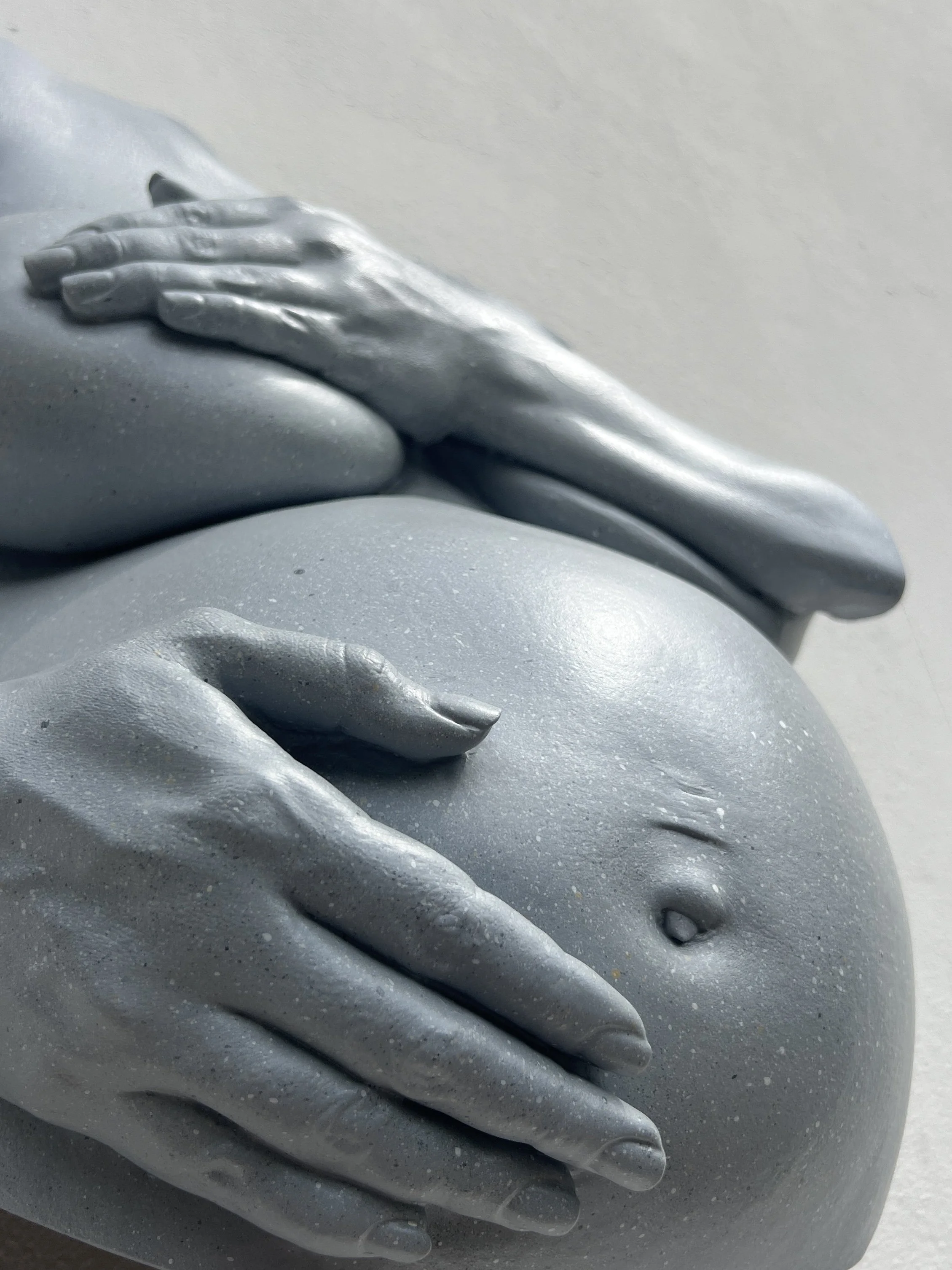 Close-up van lichtgrijs beeld van zwangere buik met borsten en handen, handgemaakt van hars met steeneffect afwerking, kunstwerk van Venus in Vorm, details van navel, zwangerschapsstriemen en handen zichtbaar.