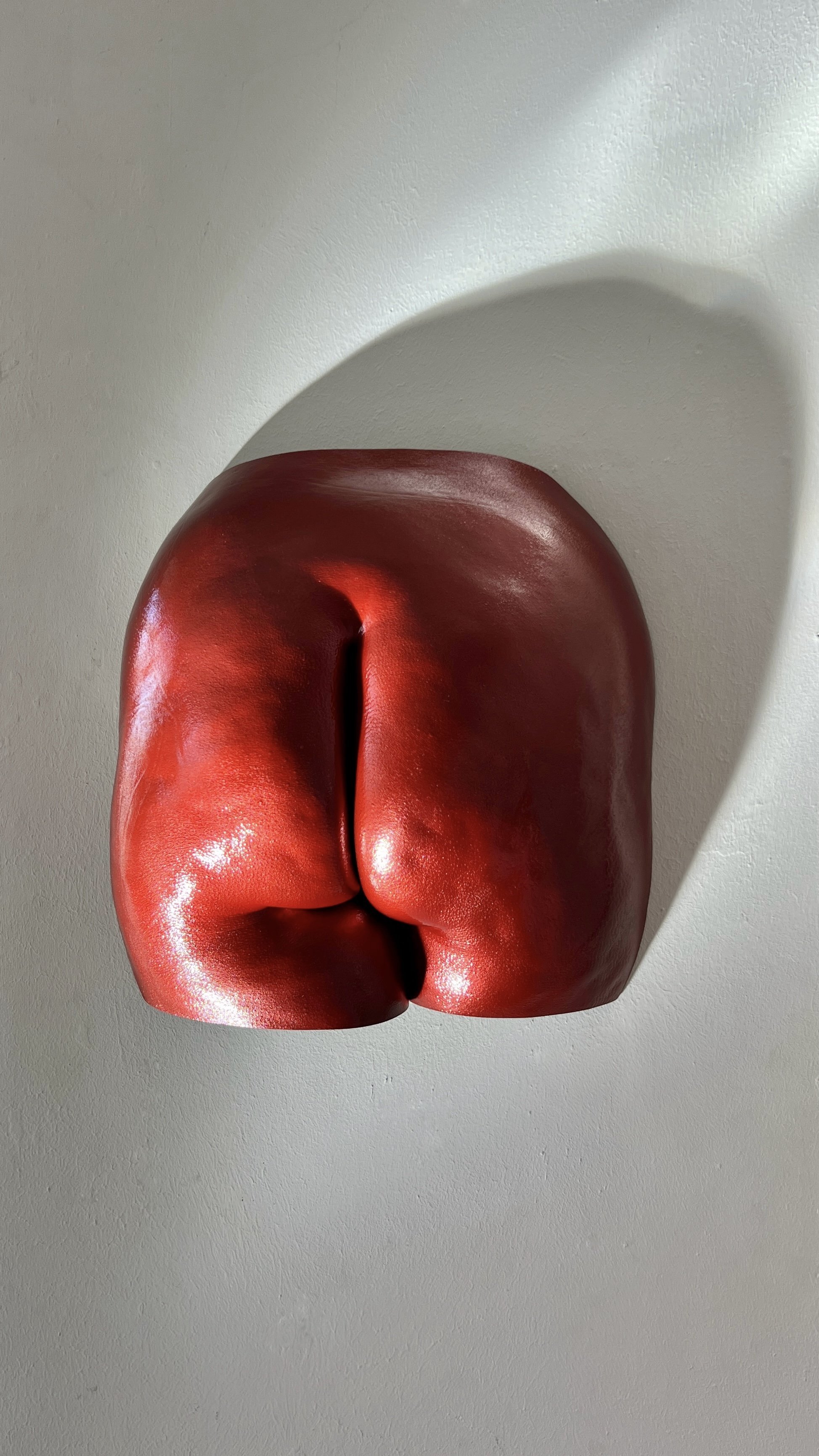 Rood metallic beeld van billen met zichtbare cellulitis en huidstructuur, handgemaakt van hars, kunstwerk van Venus in Vorm, gefotografeerd in het bodycasting atelier met spotlight die schaduw op de muur creëert.
