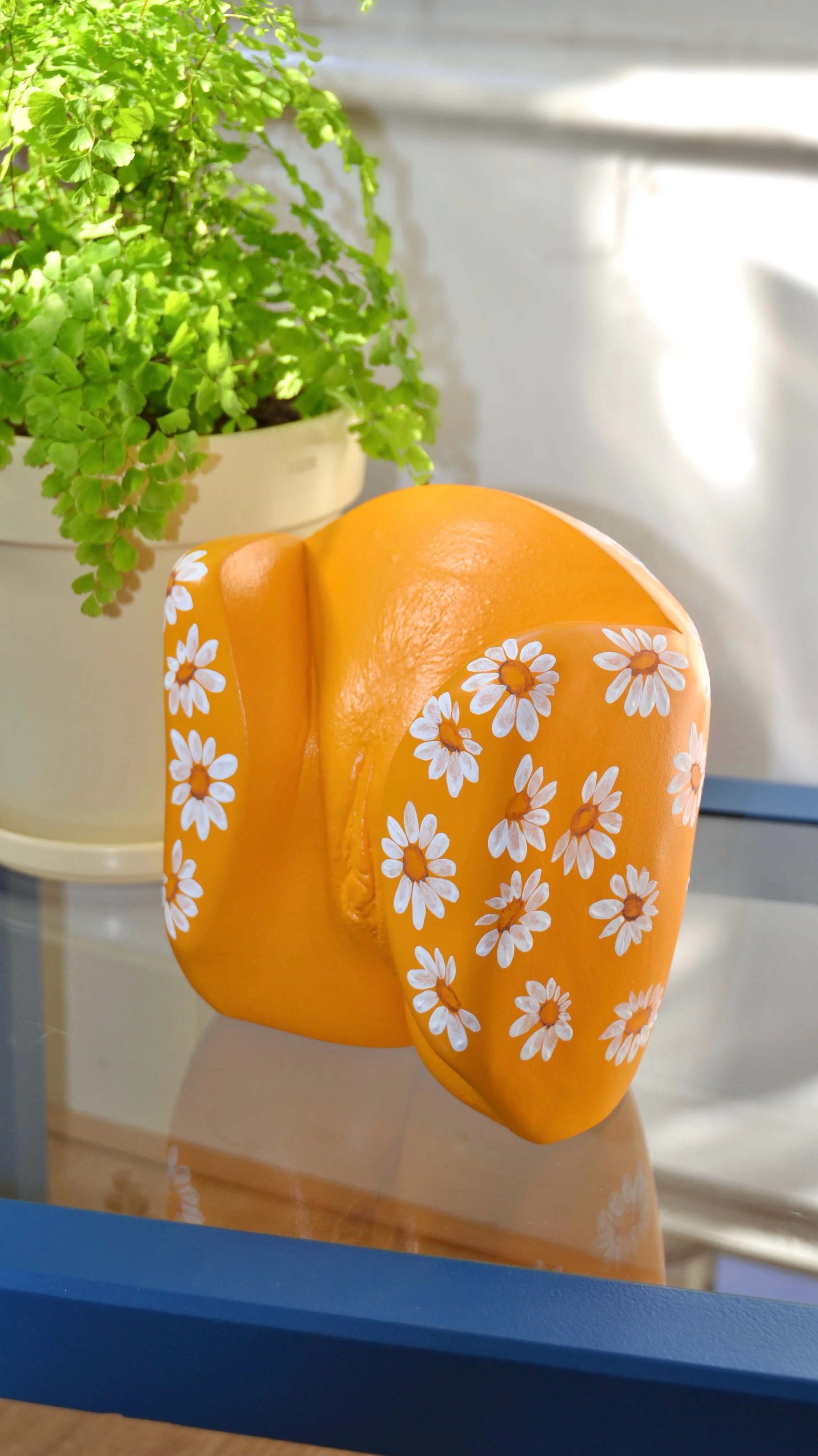 Geel vulva-beeld met handgeschilderde witte madeliefjes, handgemaakt van gips, kunstwerk van Venus in Vorm, geplaatst op een glazen tafeltje met een lichtgroene plant (Venushaar) op de achtergrond.