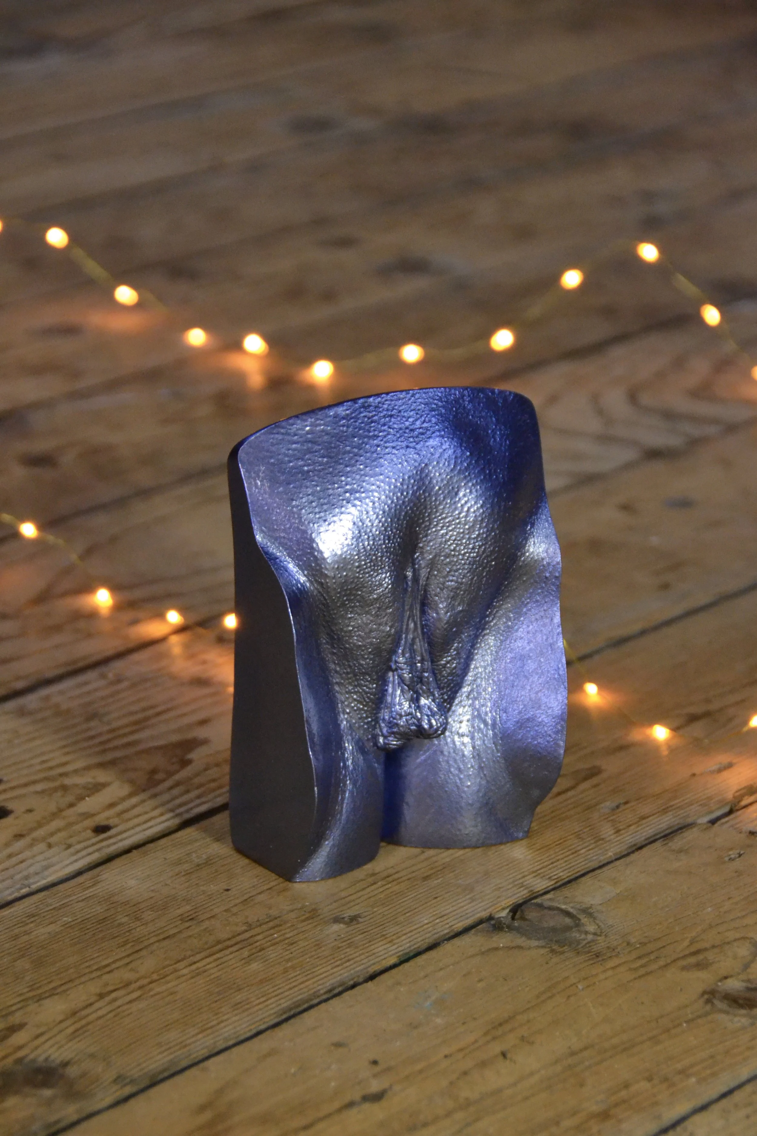 Metallic donkerblauw vulva-beeld, handgemaakt van gips, geplaatst op houten vloer met sfeervolle gele lampjes op de achtergrond.