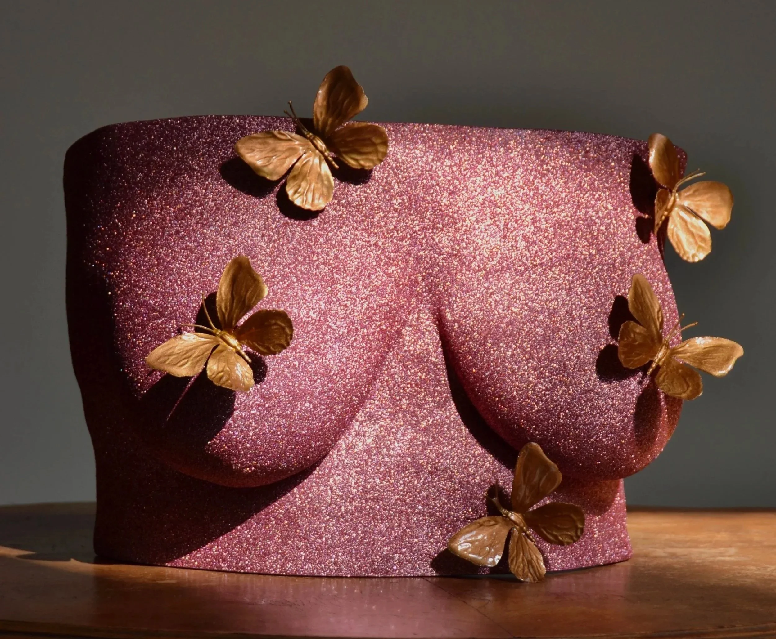 Body casting borstsculptuur met rose gold glitters en vijf decoratieve gouden vlinderornamenten, kunstwerk van Venus in Vorm, handgemaakt van hars, gefotografeerd op houten tafel in zonlicht.