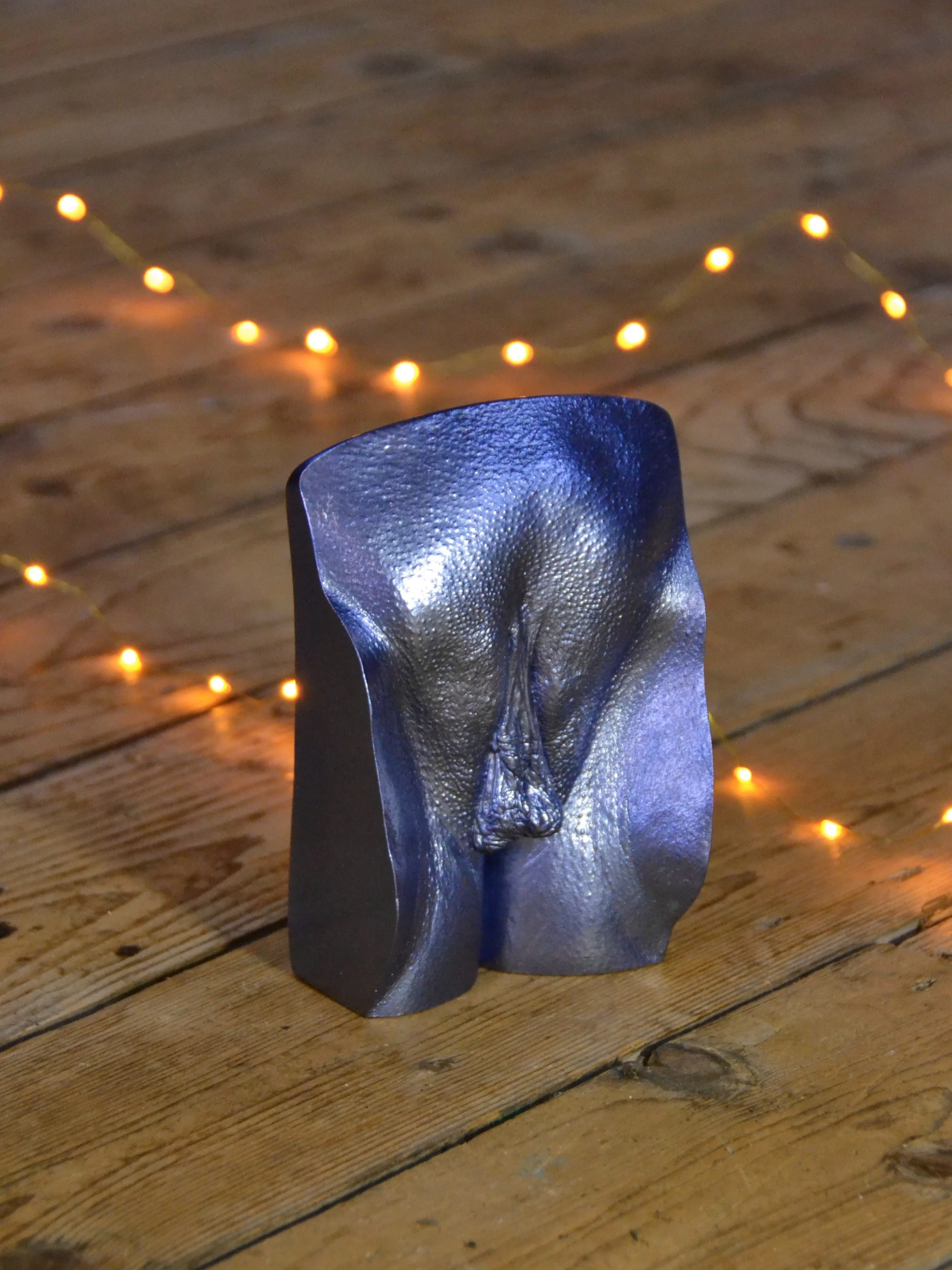 Metallic donkerblauw vulva-beeld, handgemaakt van gips, geplaatst op houten vloer met sfeervolle gele lampjes op de achtergrond.