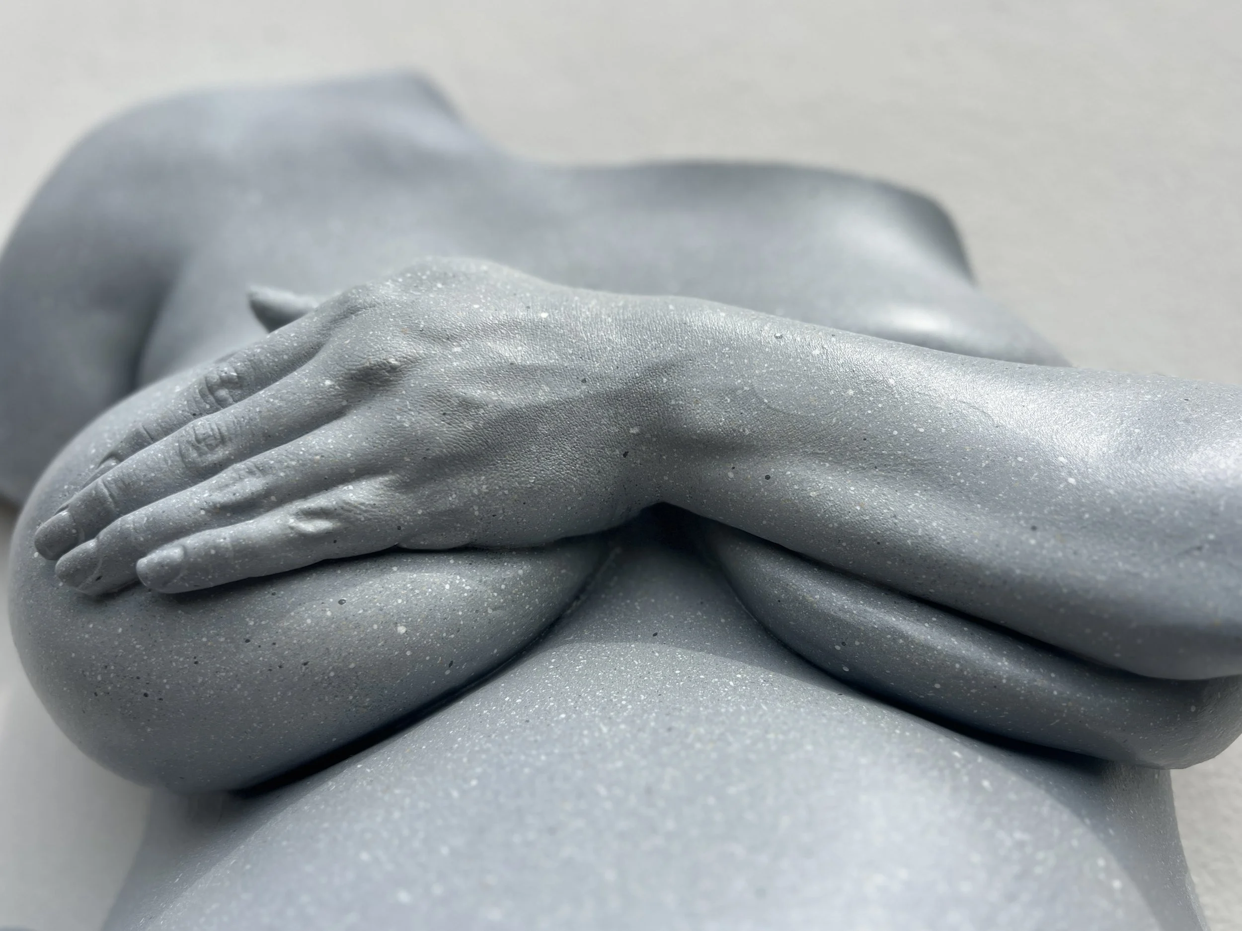 Close-up van lichtgrijs beeld van zwangere buik met borsten bedekt door hand en onderarm, handgemaakt van hars met steeneffect afwerking, details van hand en tatoeage op onderarm zichtbaar, kunstwerk van Venus in Vorm.