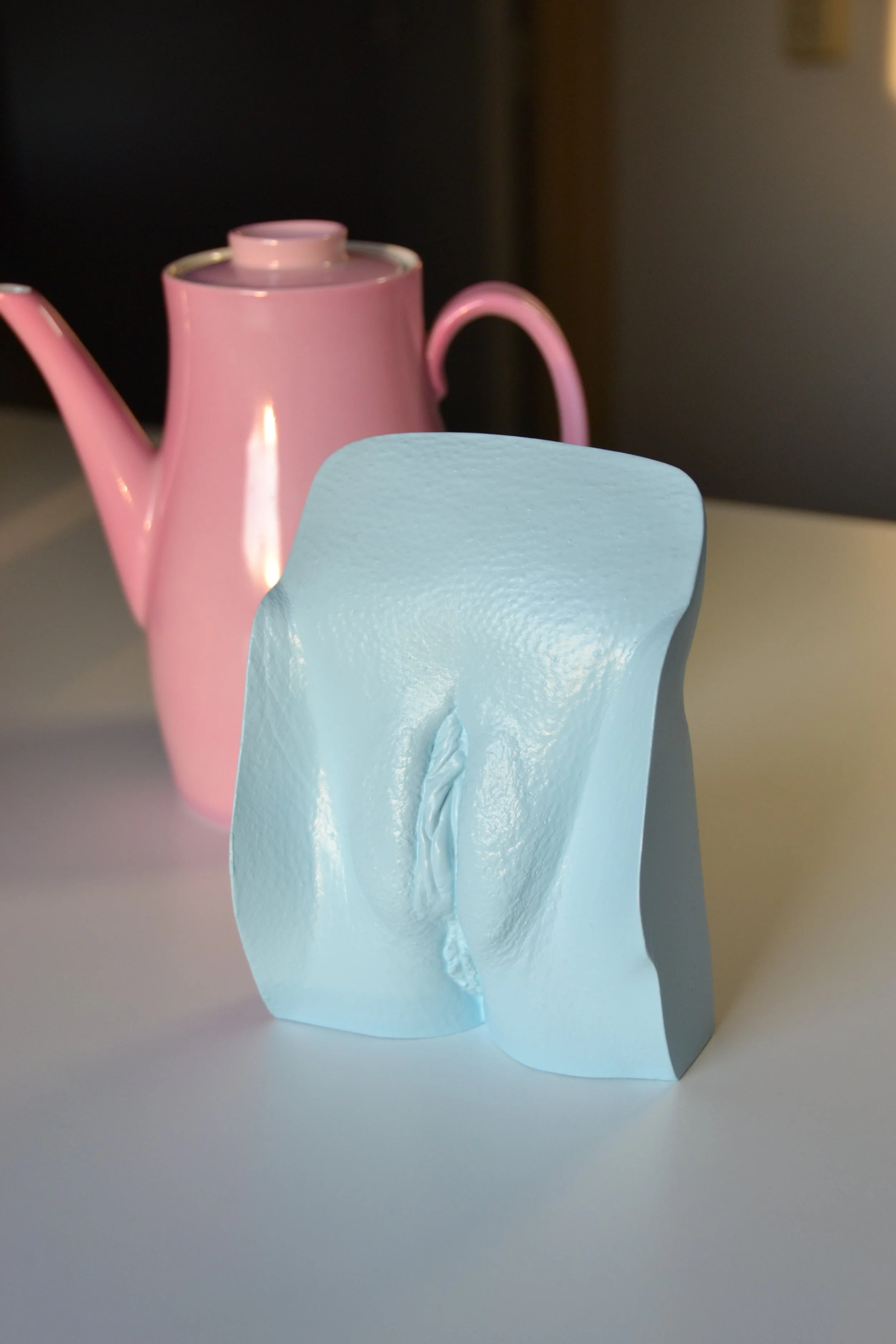 Lichtblauw vulva-beeld, handgemaakt van gips, geplaatst op een witte tafel met een roze theepot op de achtergrond, in atelier Venus in Vorm.