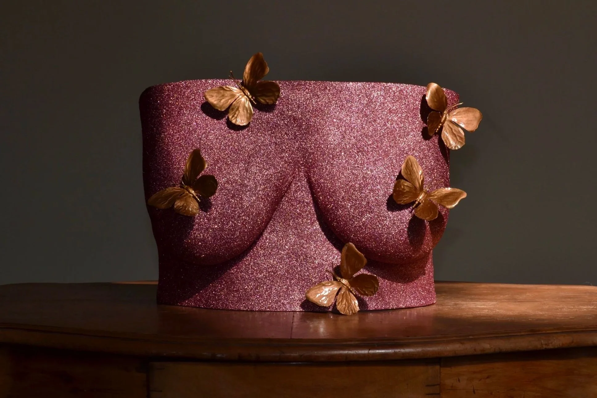 Vooraanzicht van body casting borstsculptuur met rose gold glitters en vijf decoratieve gouden vlinderornamenten, kunstwerk van Venus in Vorm, handgemaakt van hars, gefotografeerd op houten tafel in zonlicht.
