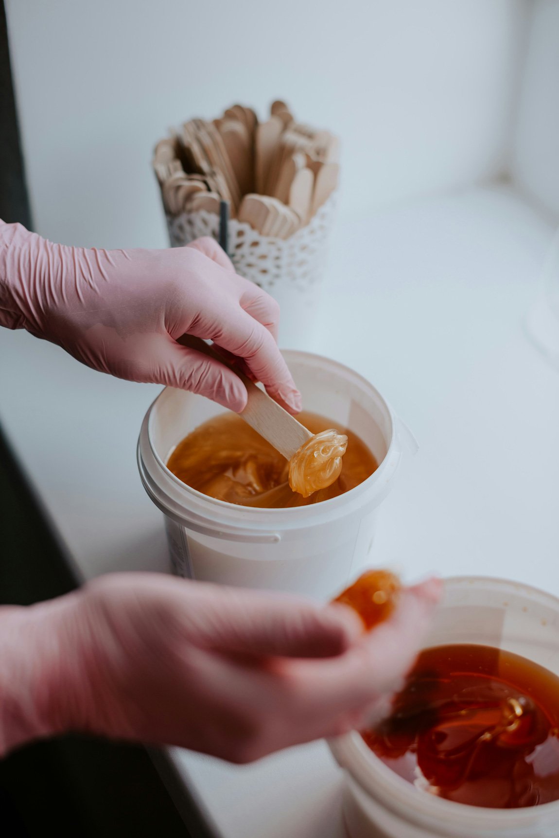 Close-up of gloved hands preparing soft wax with spatula from container, part of professional waxing service prep