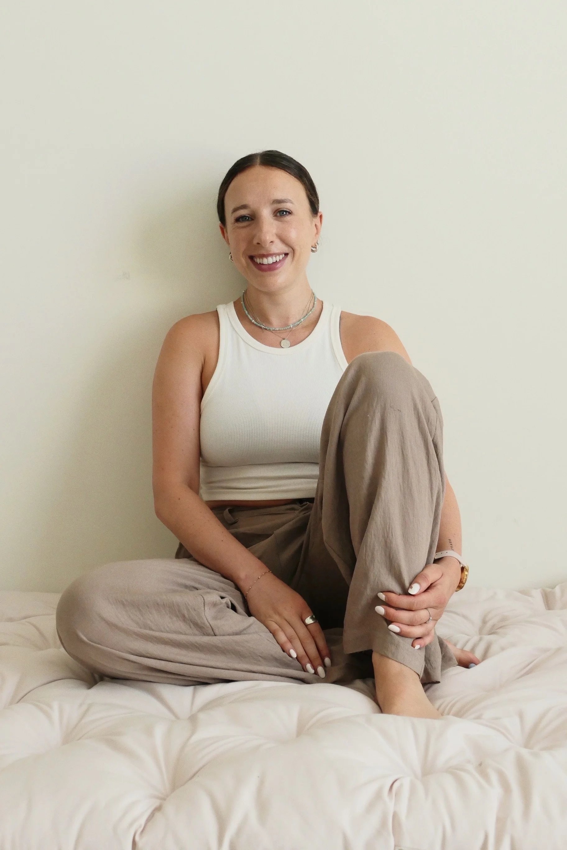 A smiling woman sitting cross-legged on a beige bed with a light-colored wall behind her. She is wearing a white tank top, beige pants, and jewelry, including necklaces, rings, and a watch.