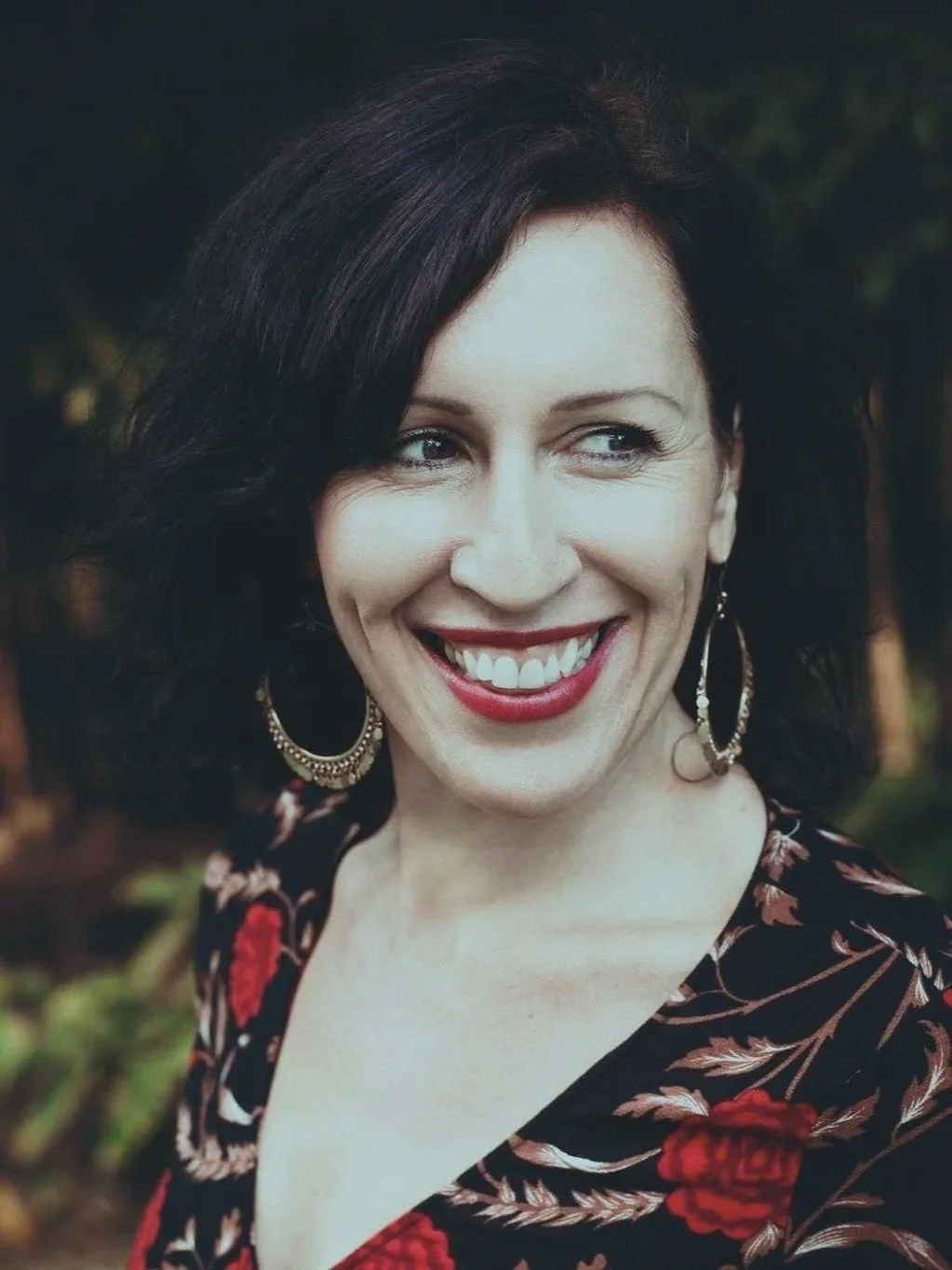 Shae Allen founder of ALSHA Work with dark hair styled in loose waves, smiling and wearing large hoop earrings, a red lipstick, and a black blouse with red floral patterns, outdoors with blurred trees in the background.