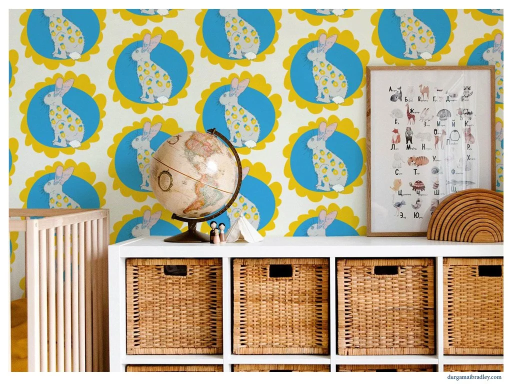 Child's room with a wallpaper featuring rabbits inside yellow and blue scalloped circles. On white furniture, there is a globe, small figurines, a framed poster with animals and alphabet, and wooden rainbow stacking toys.