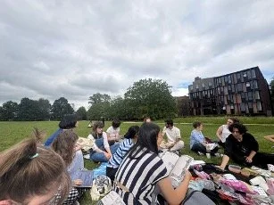 A shot of part of a group of students sitting in a park, reading our magazine, The WordHoard. The sky is cloudy and they are having a picnic.