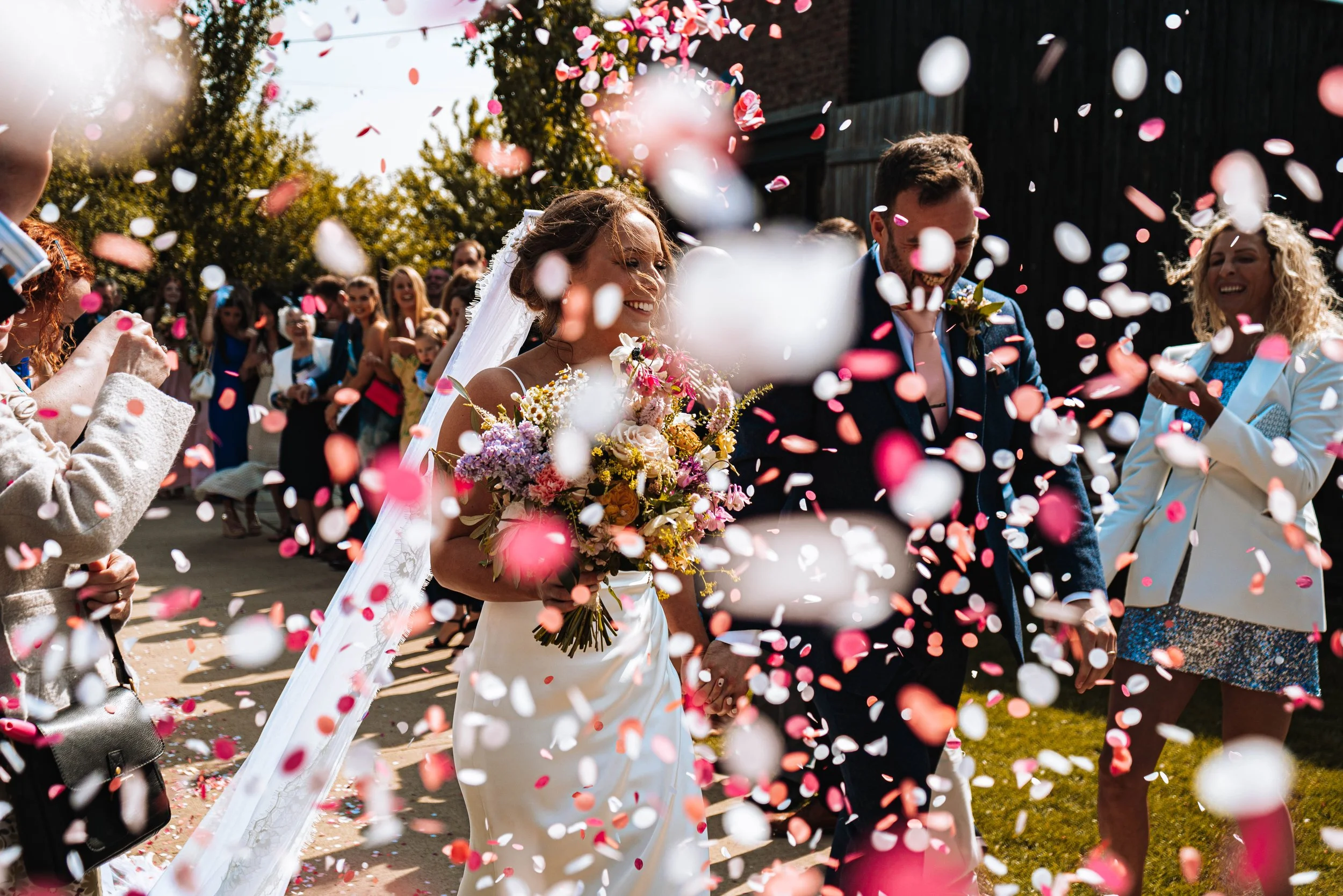 Bride and groom surrounded by guests throwing pink and white confetti outdoors on a sunny day.