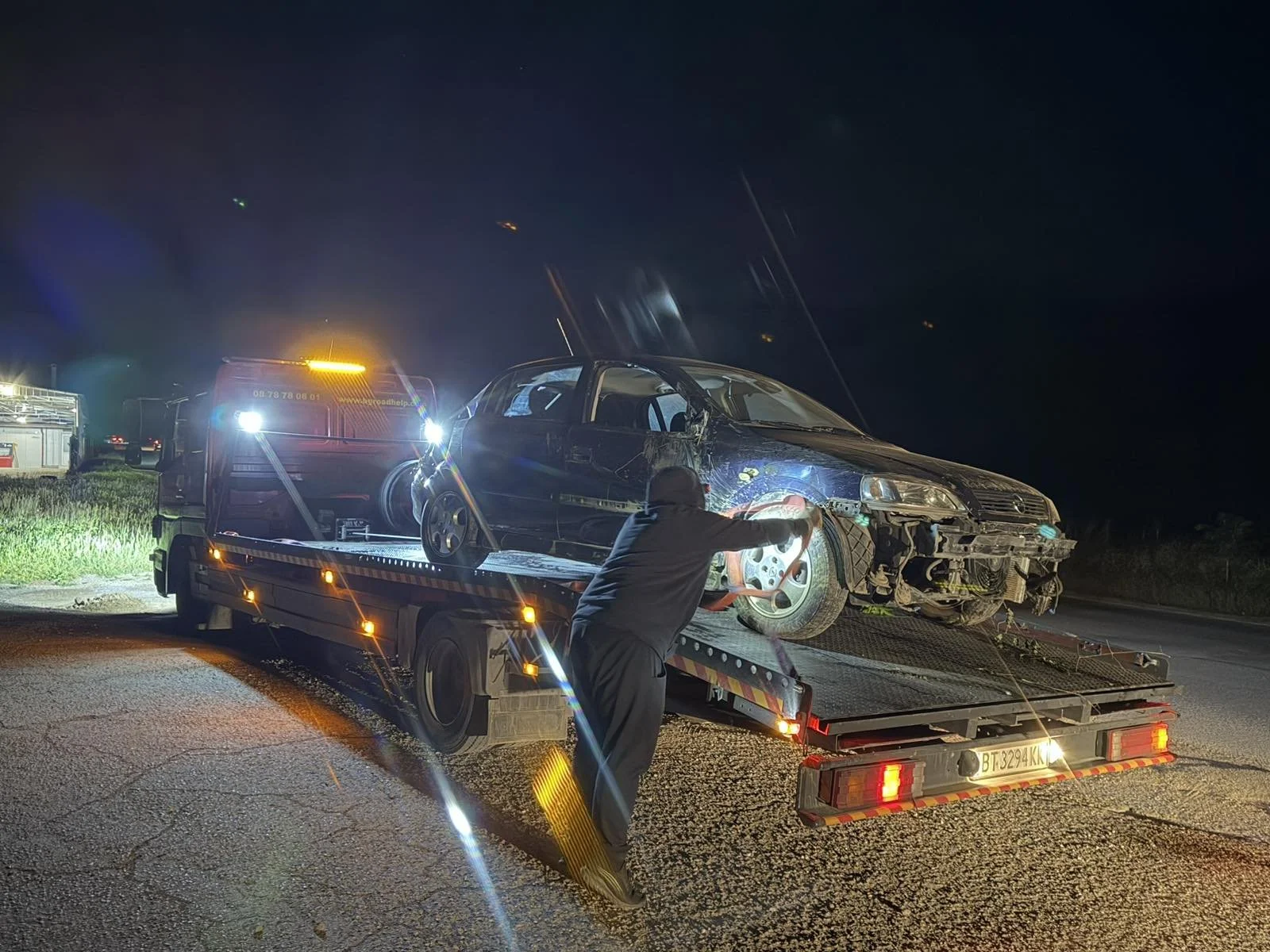 Nighttime roadside scene showing a damaged black car on a flatbed tow truck. A person is working on the car, using a tool near the front wheel. Emergency or repair vehicle with flashing lights is present in the background.