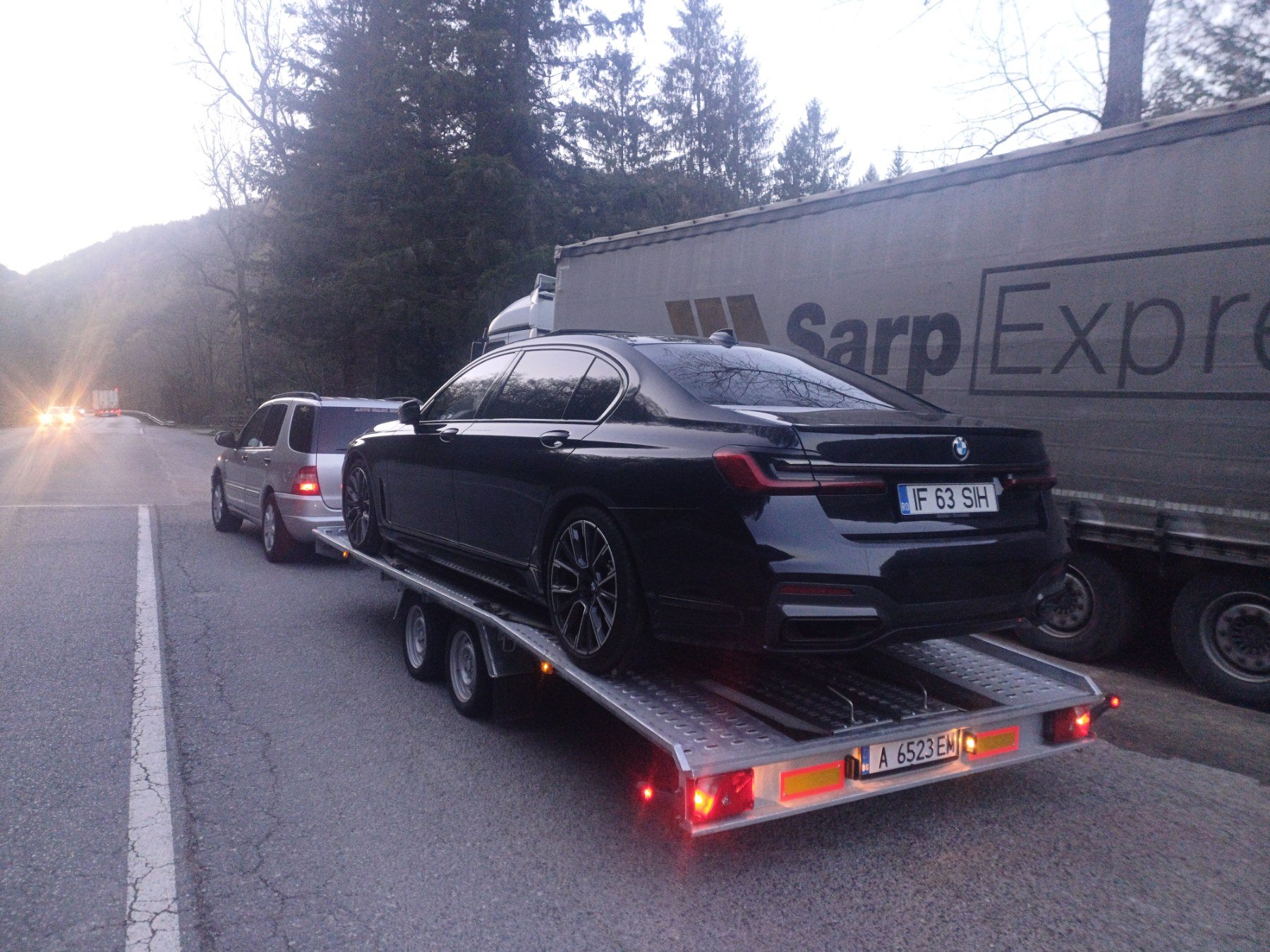 A car being towed on a trailer behind a gray vehicle on a roadside near a truck, with trees and a hill in the background.