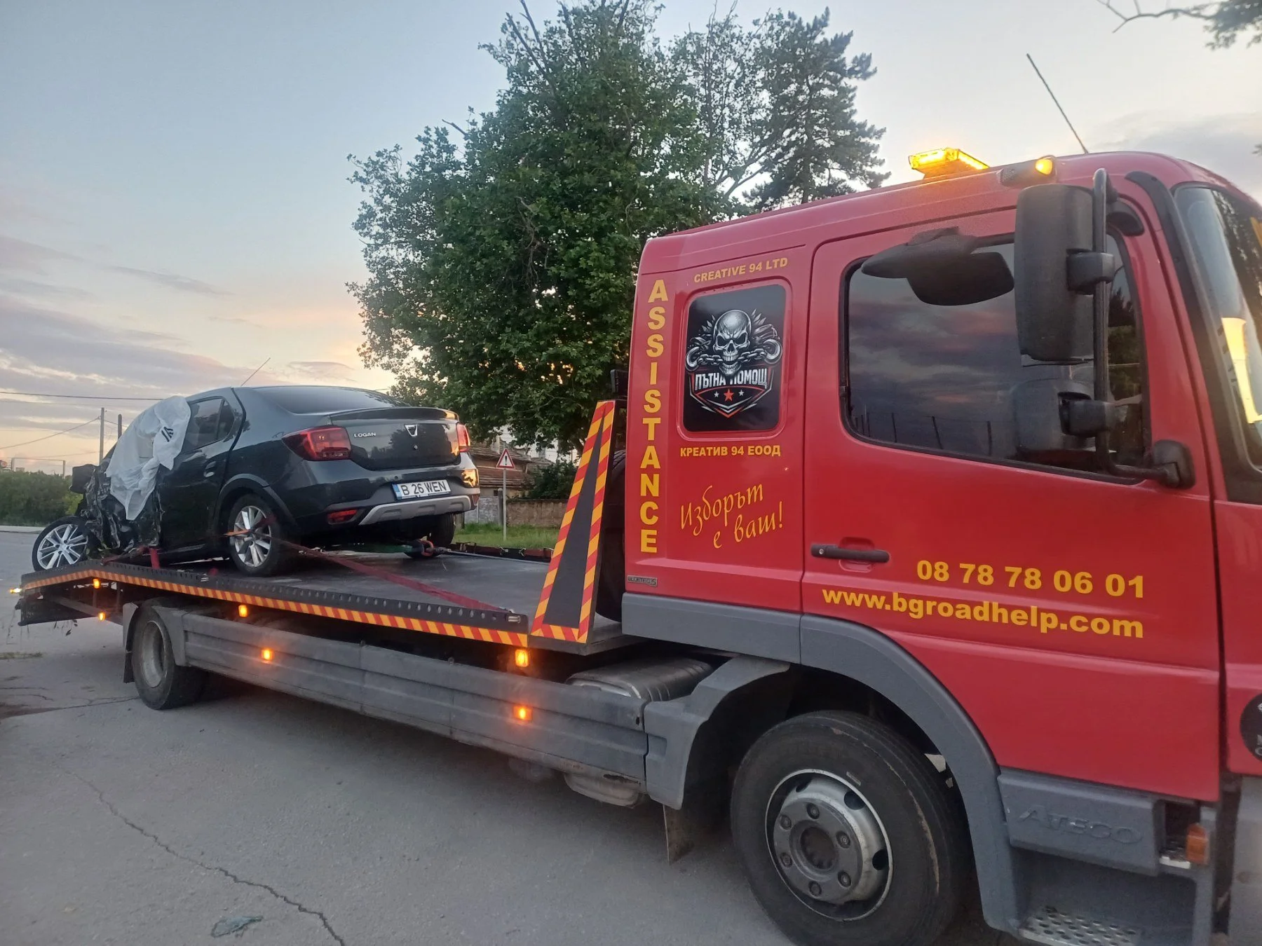 A red tow truck with decorations and text, carrying a black car with damage to the front and side, on a flatbed. The sky is partly cloudy and there are trees in the background.