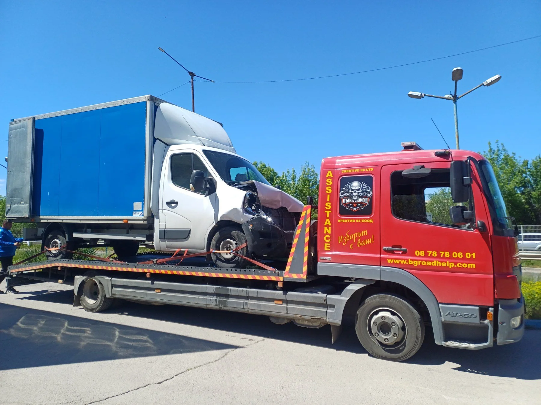 Red tow truck transporting a damaged white delivery truck on a flatbed. The damage is seen on the front part of the white truck. The sky is clear with some trees in the background.