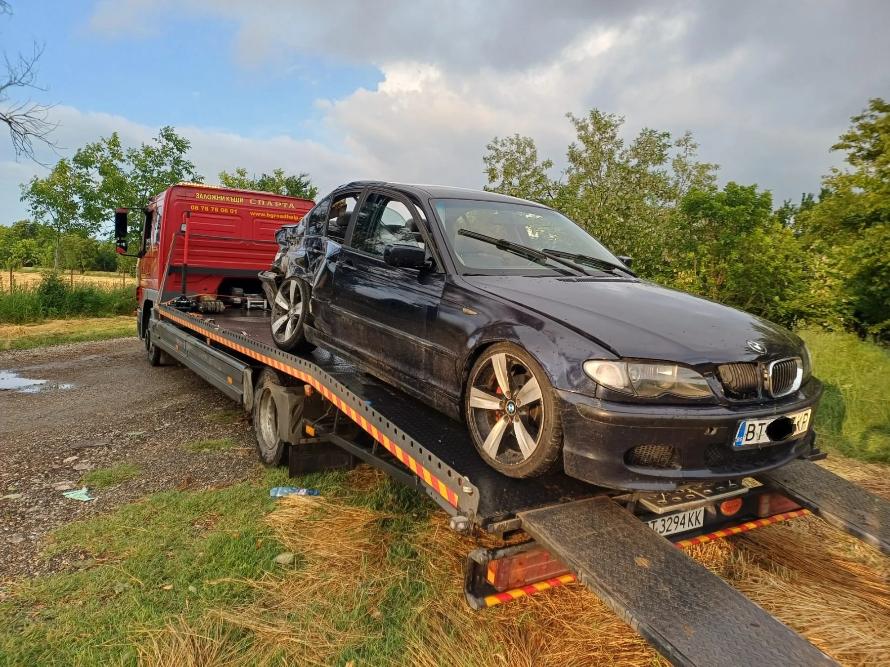Black damaged BMW sedan being transported on a tow truck with a red truck behind it, parked on a grassy roadside with trees and cloudy sky in the background.