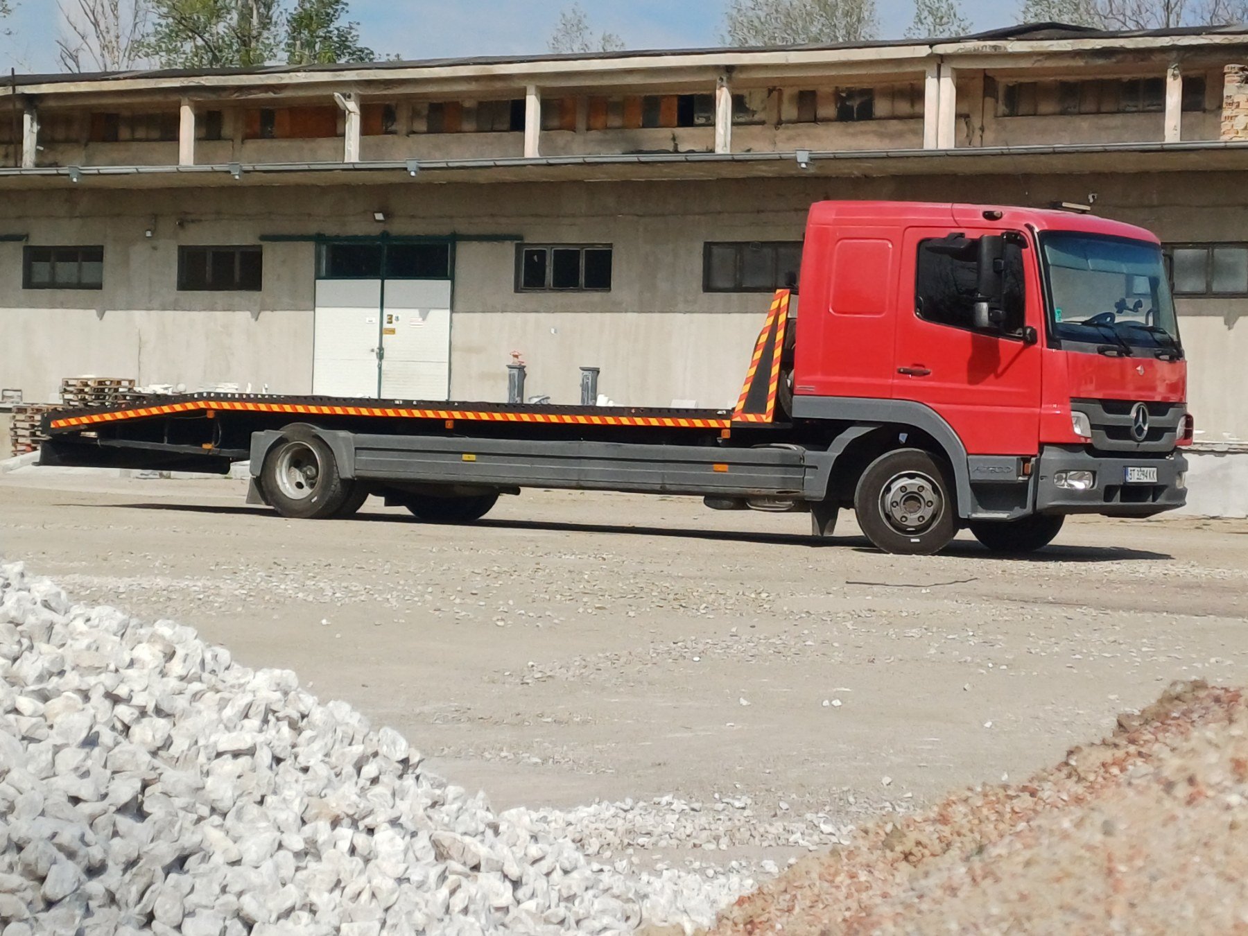 Red flatbed truck parked on gravel construction site in front of unfinished building.