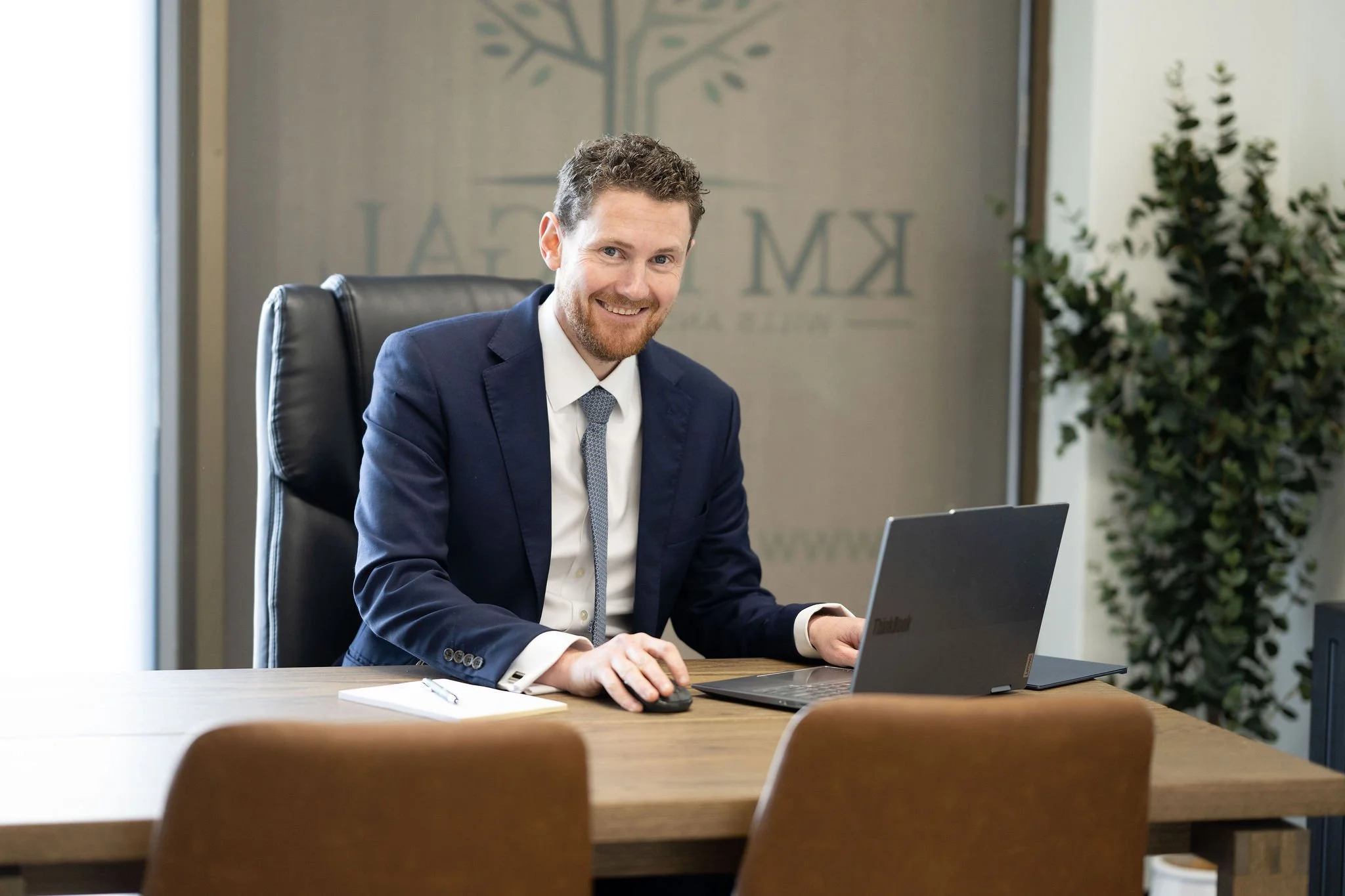 A smiling man in a navy blue suit and tie sitting at a wooden conference table with a laptop and notepad, in an office with a logo on the wall and green plants.