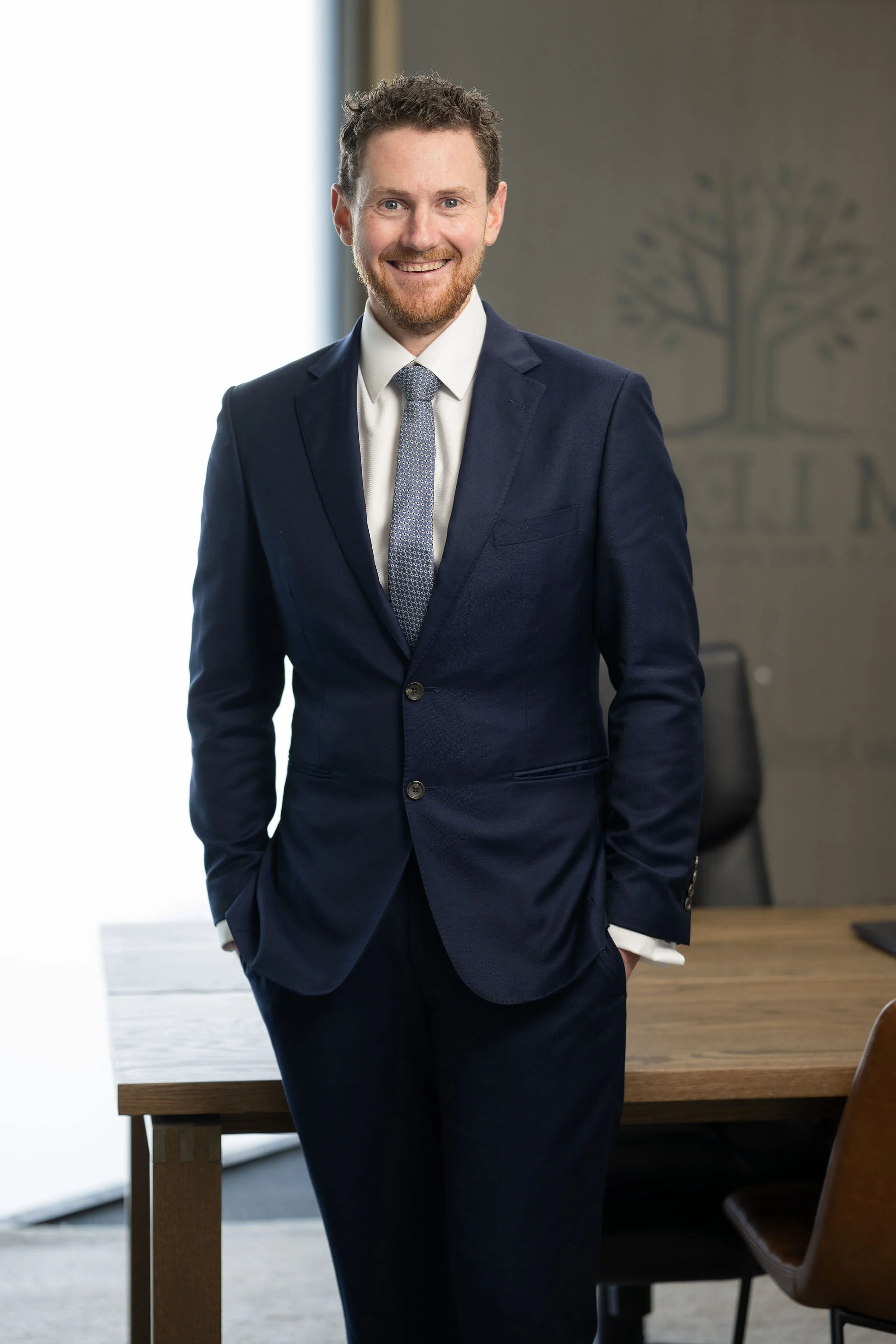 A man with light skin, curly brown hair, and a beard, wearing a dark blue suit, white shirt, and patterned tie, stands confidently in a modern office with a large window and a wall with a tree logo in the background.