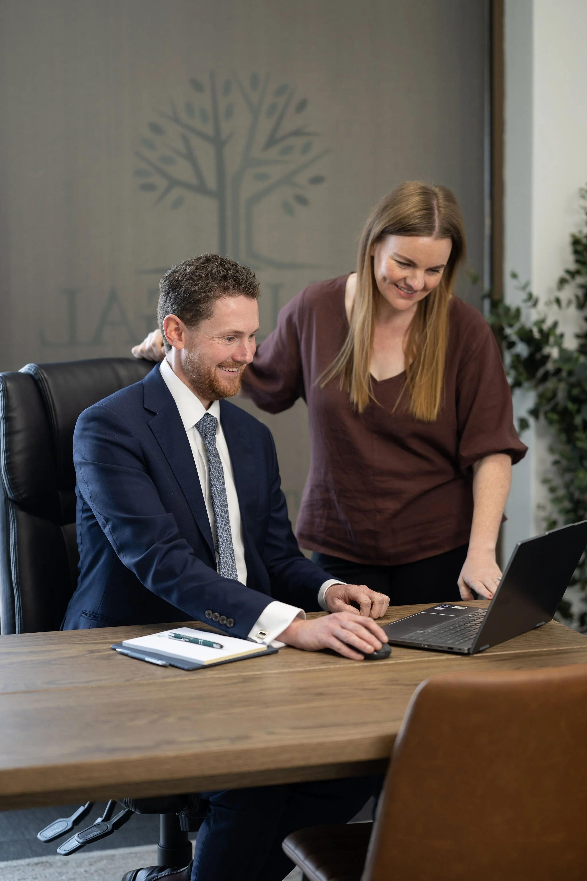 A man in a navy suit and a woman in a brown top looking at a laptop on a wooden table in a meeting room.
