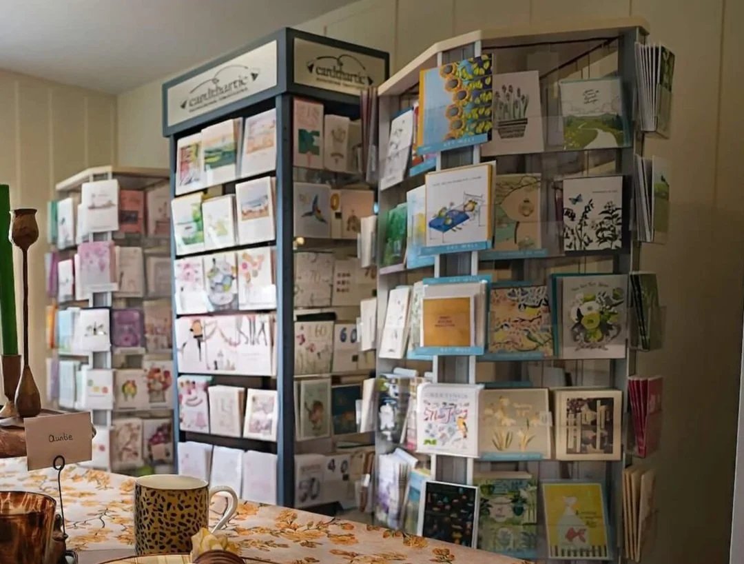 Multiple greeting cards displayed on photo display racks in a store. A table with a floral tablecloth, a mug, a small sign, and a candle are visible in the foreground.