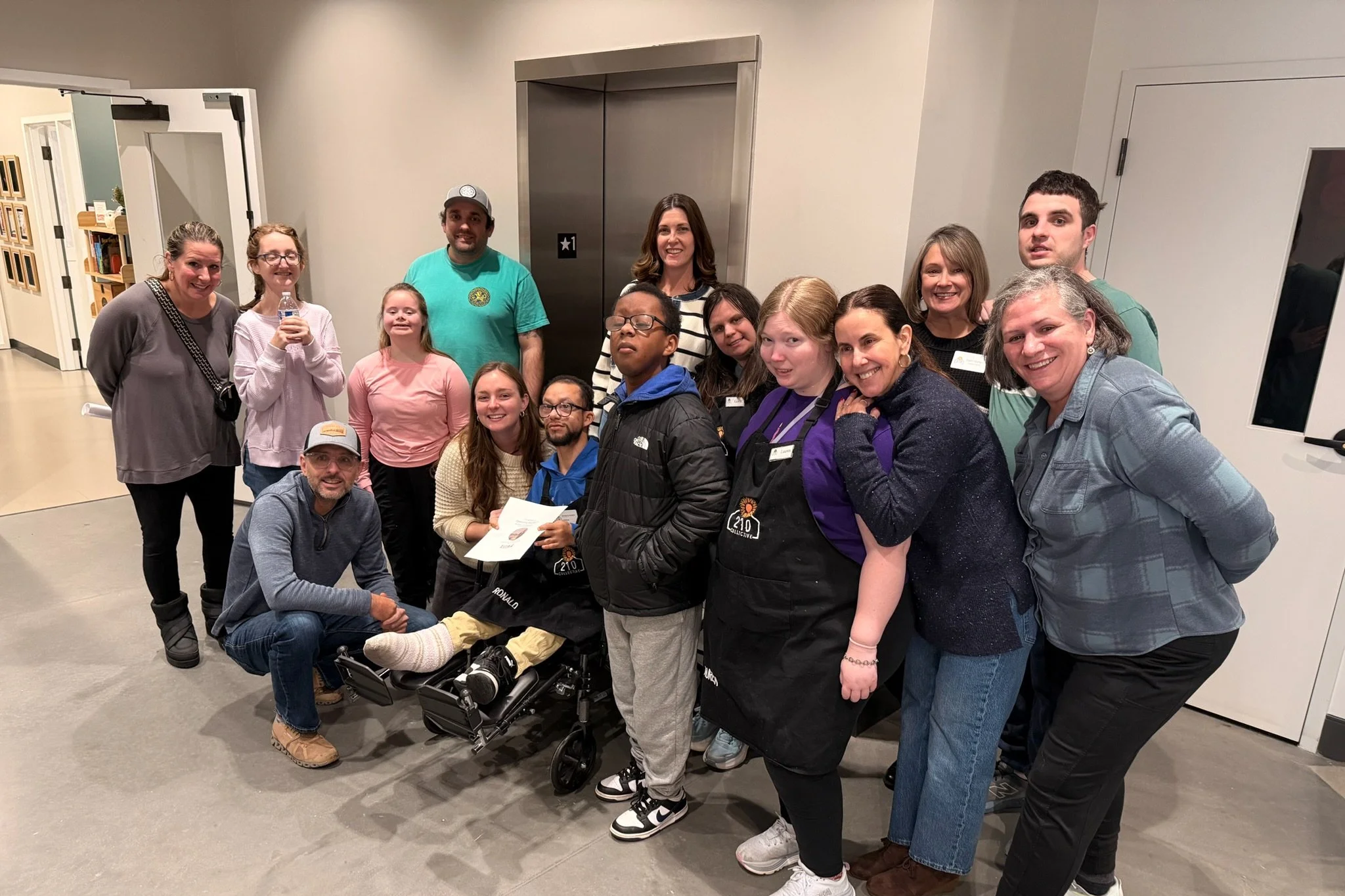A group of fifteen people, including men, women, and children, gathered together indoors near an elevator. They are smiling and posing for a group photo, with some standing and others kneeling. The background shows a beige wall, an elevator, and a do