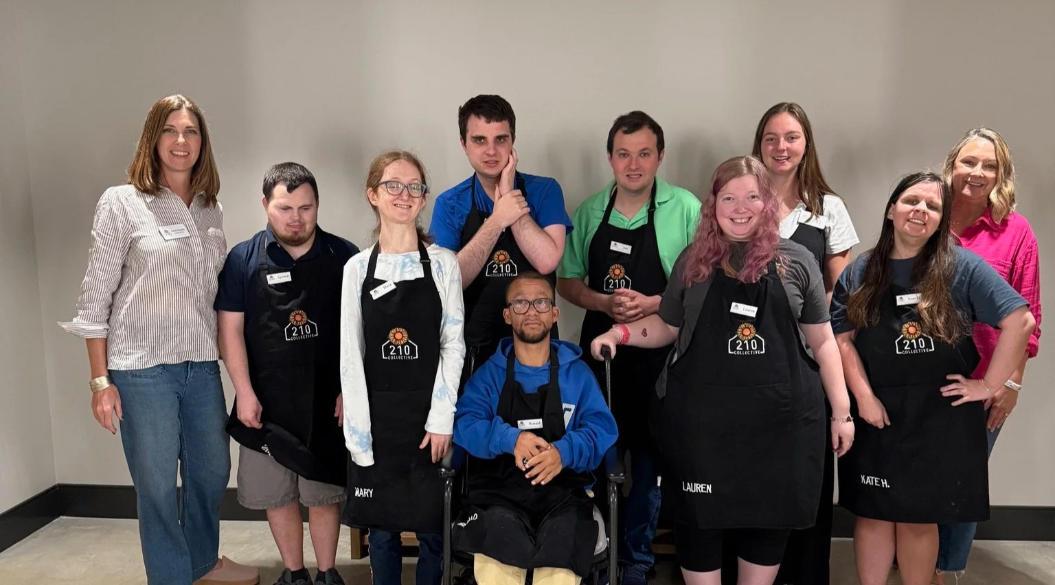 Group of people standing and sitting together, some wearing aprons with '210 Collective' logo, in a room with plain wall.