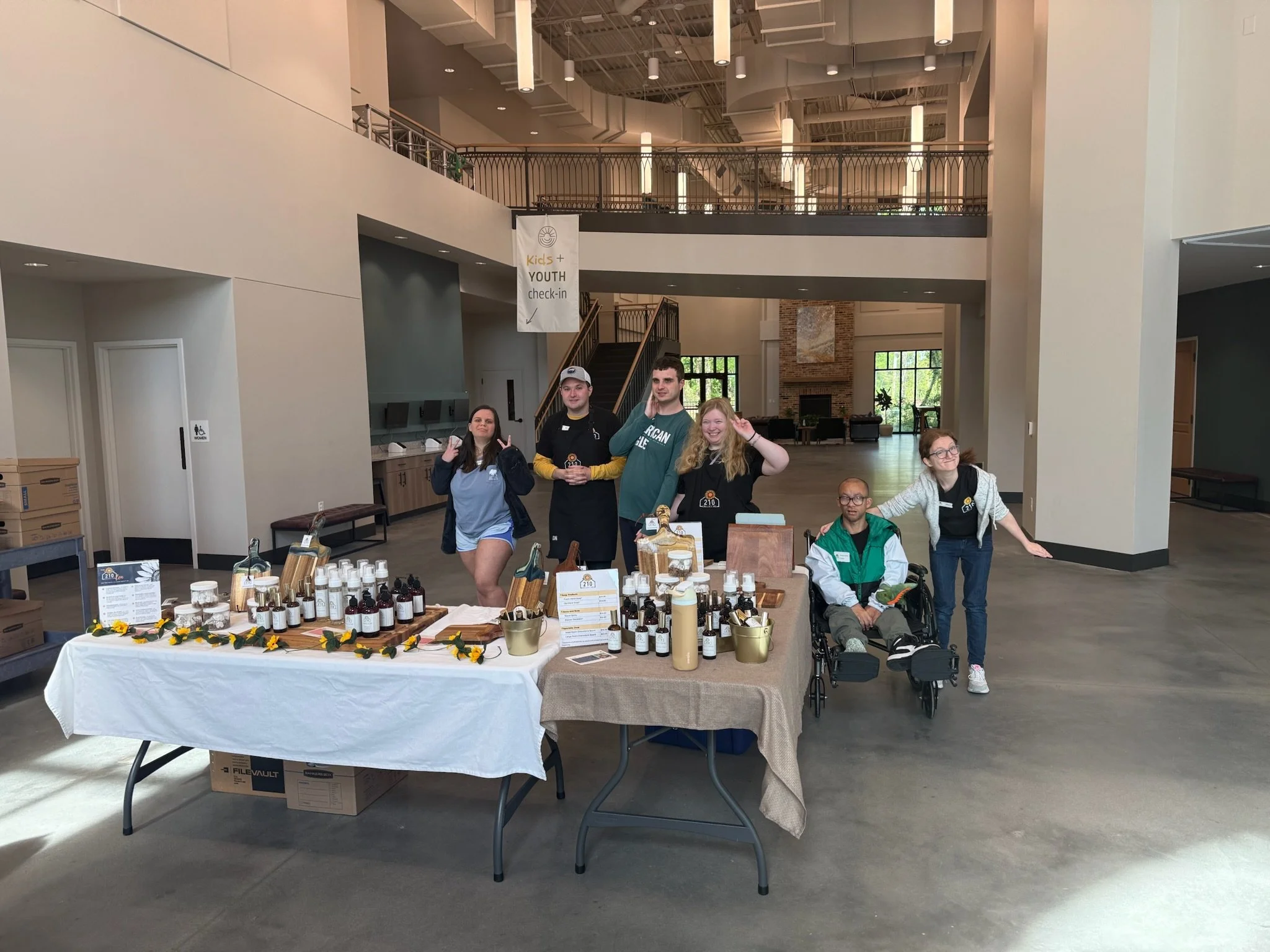 Group of five young people standing behind a table displaying various bottles and products at an indoor event or market, with a two-story interior including a staircase, a balcony, and large windows in the background.