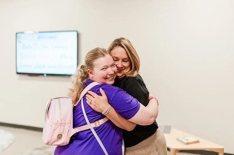 A girl in a purple shirt hugging a woman in a black shirt
