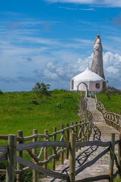 Caminho de madeira levando a uma pequena igreja branca em um campo verde, com uma estátua alta ao fundo. Céu azul com algumas nuvens.