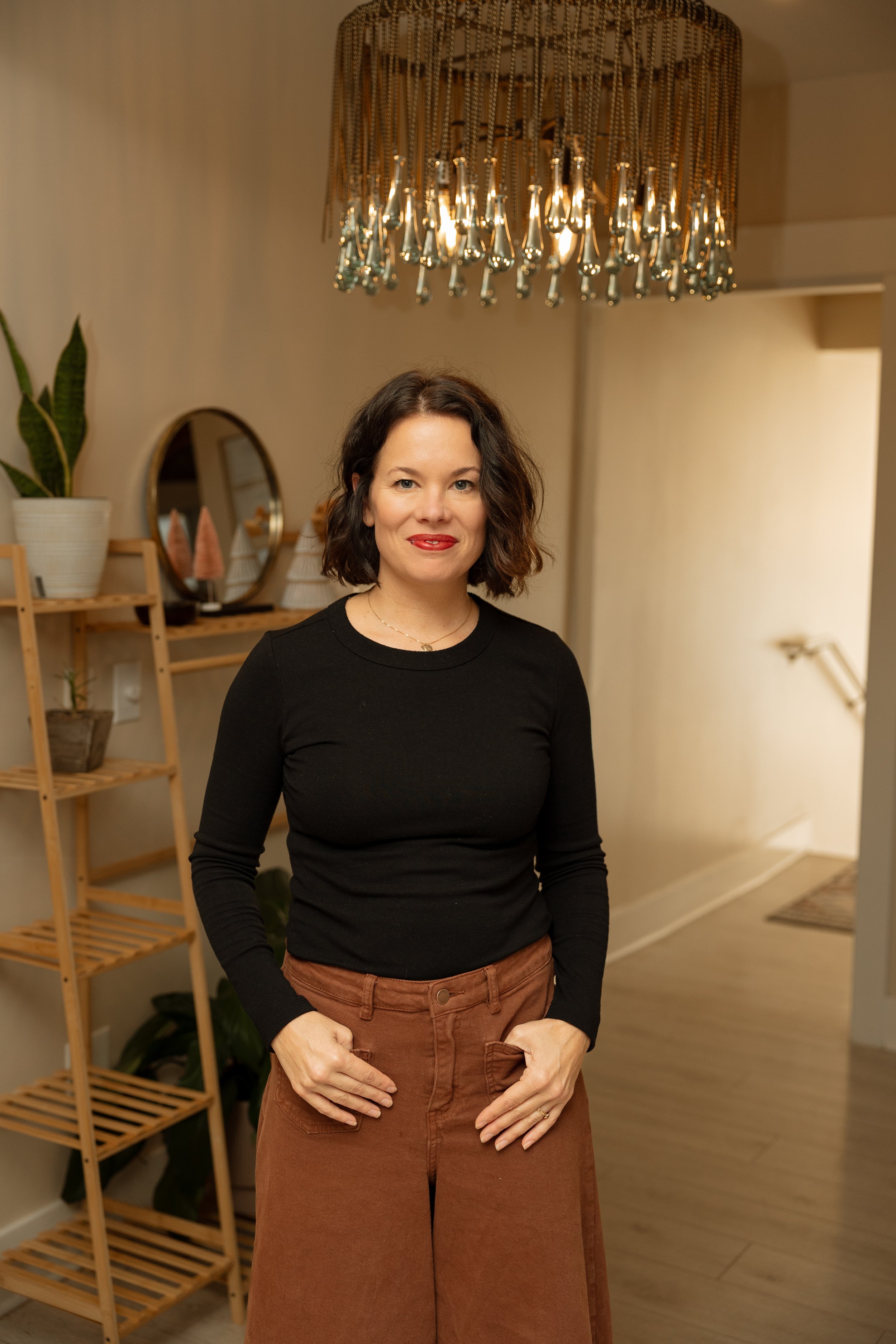 Woman sitting on the floor near a black and white wall, wearing a white blazer, beige top, light-colored pants, and brown shoes.