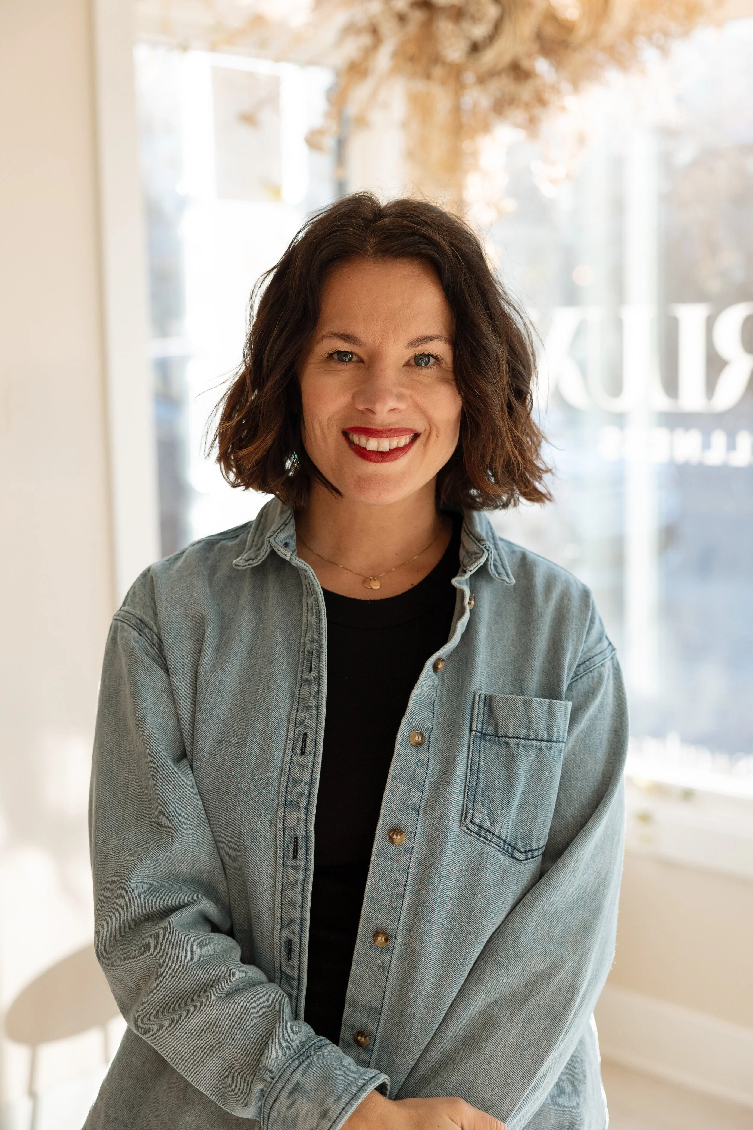Young woman with short dark hair smiling, wearing a black shirt and a light denim jacket, standing indoors near a window.