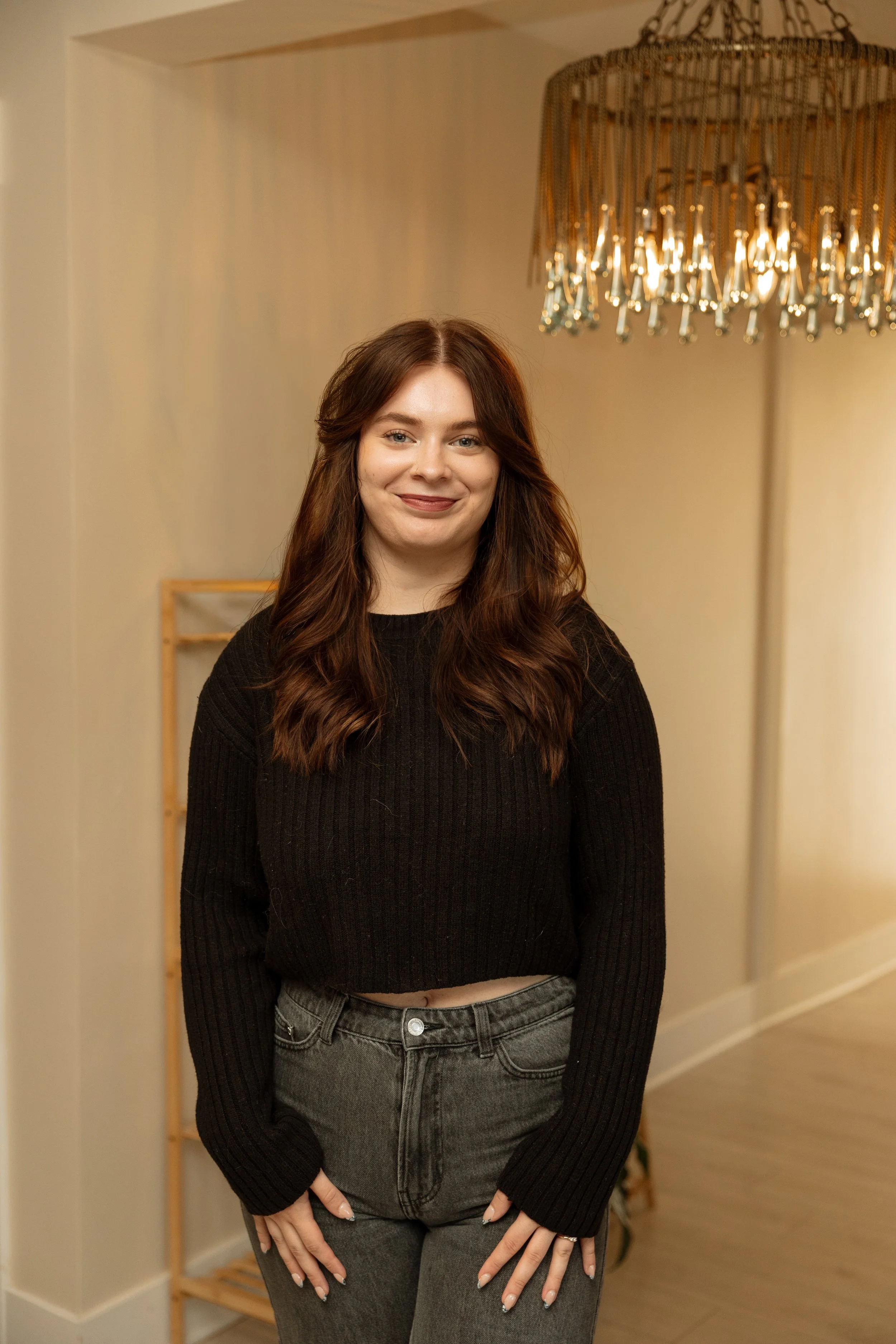 Close-up of a smiling woman with wavy dark hair, wearing a brown satin top and gold necklaces, with her eyes closed and hand resting on her head.