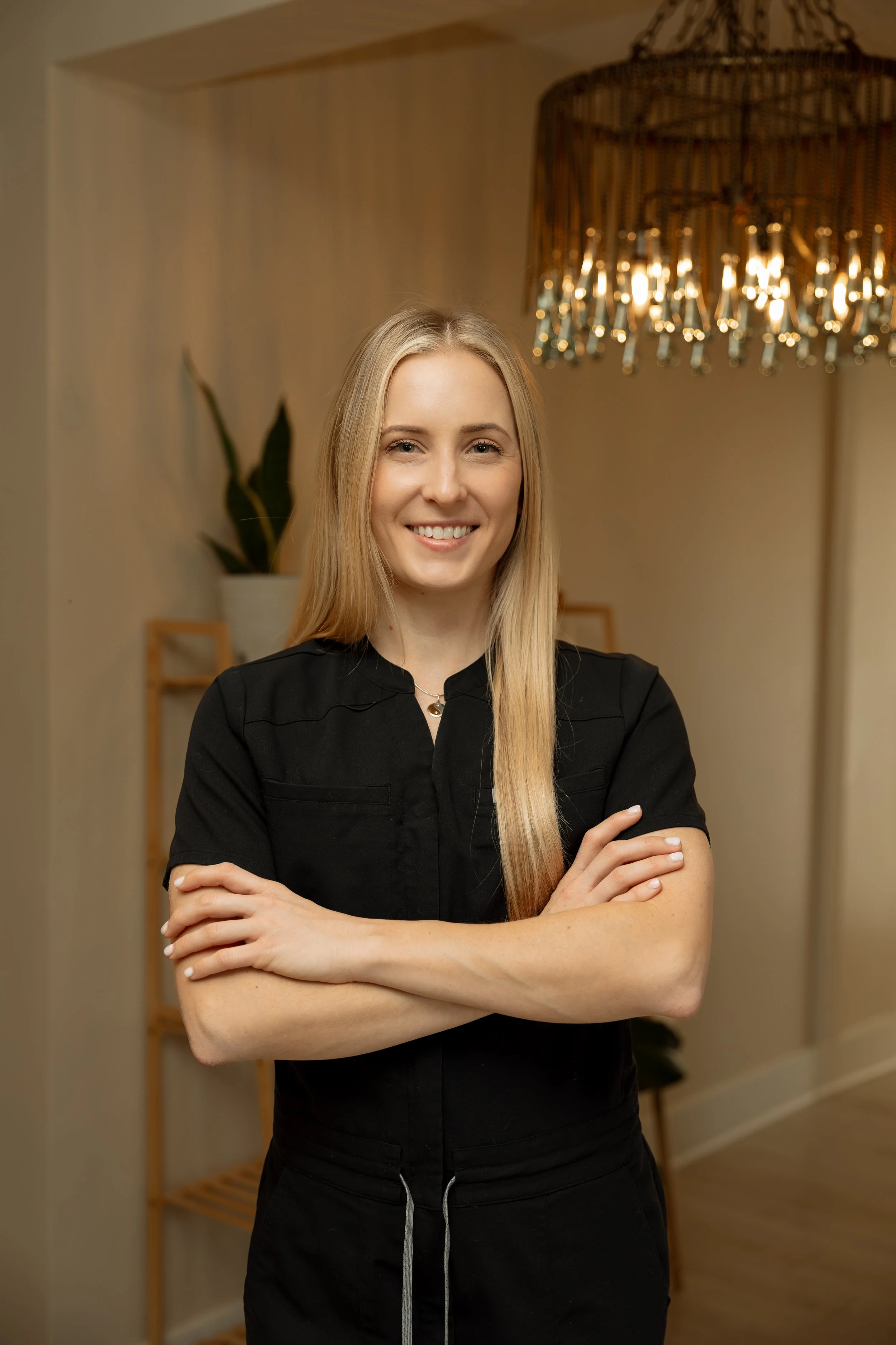 A smiling blonde woman in a black shirt with arms crossed standing in a well-lit room with a decorative chandelier and a plant in the background.