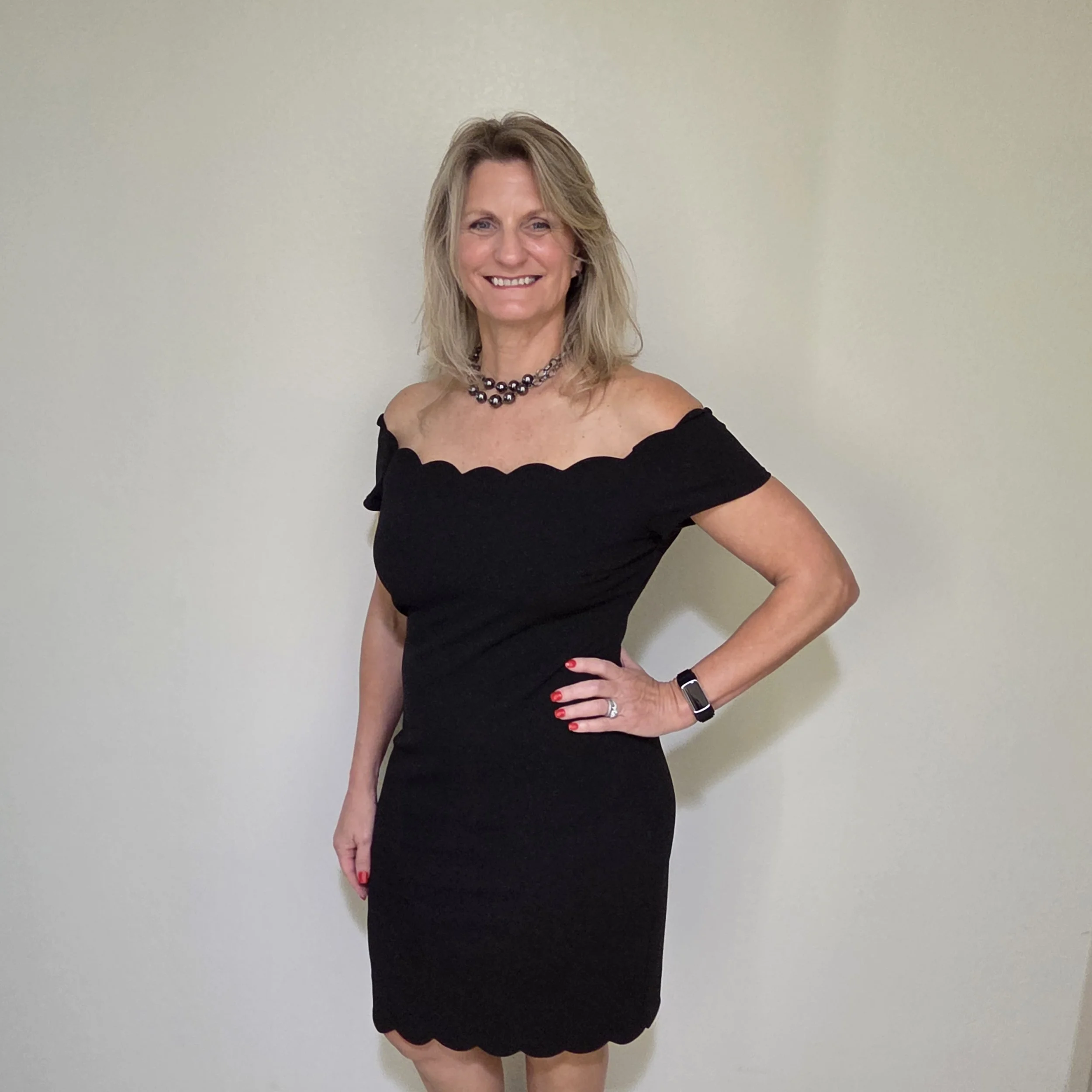 A smiling woman with shoulder-length blonde hair wearing a black off-the-shoulder dress, a pearl necklace, red nail polish, and a black wristwatch, standing against a plain light-colored wall.