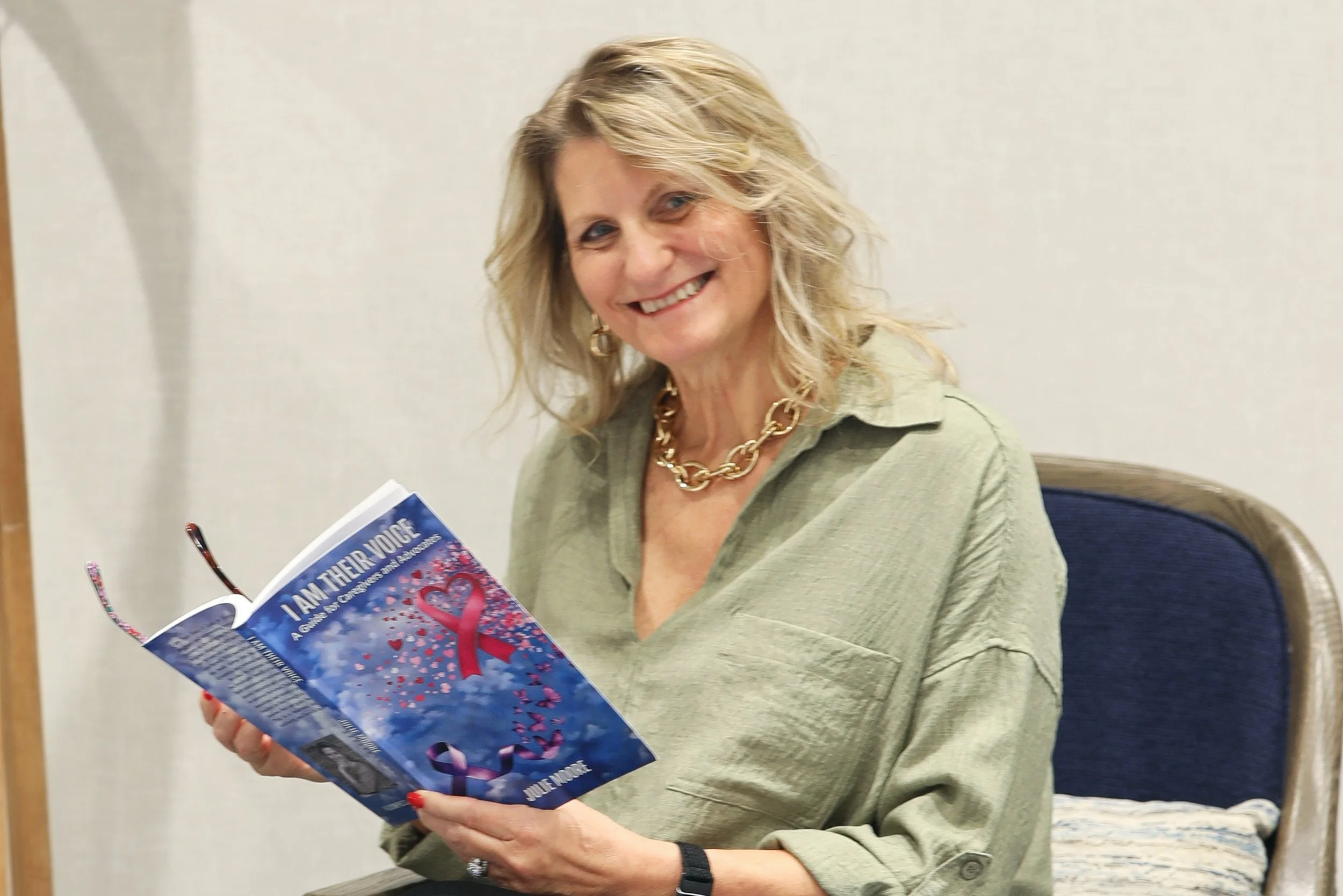 A smiling woman with blonde hair and gold jewelry sits at a table, holding a booklet titled 'I Am Their Voice' with a pink ribbon on the cover.