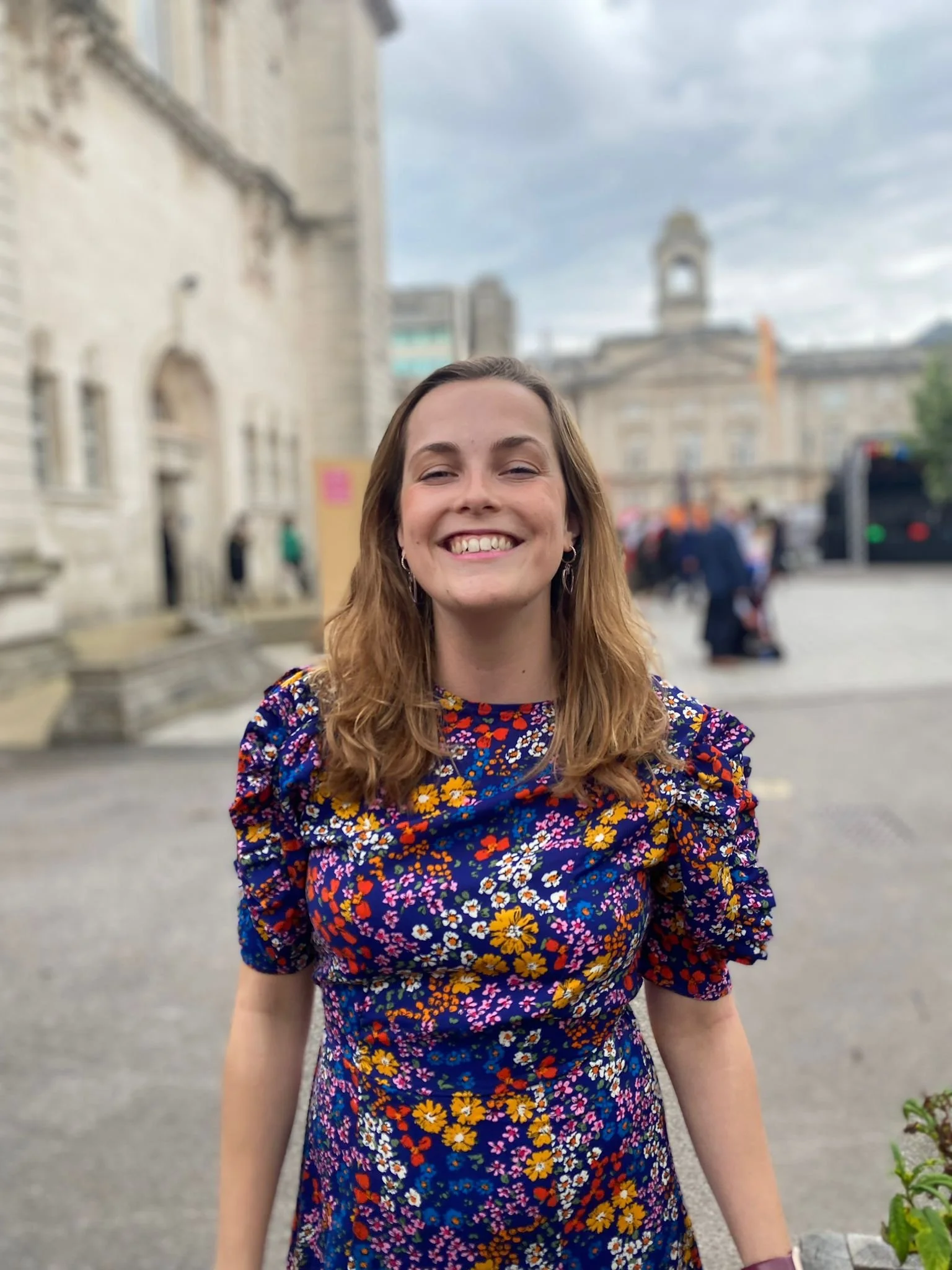 A young woman with shoulder-length light brown hair smiling outdoors in front of a historic building with a clock tower. She is wearing a colorful floral dress with puffed sleeves.