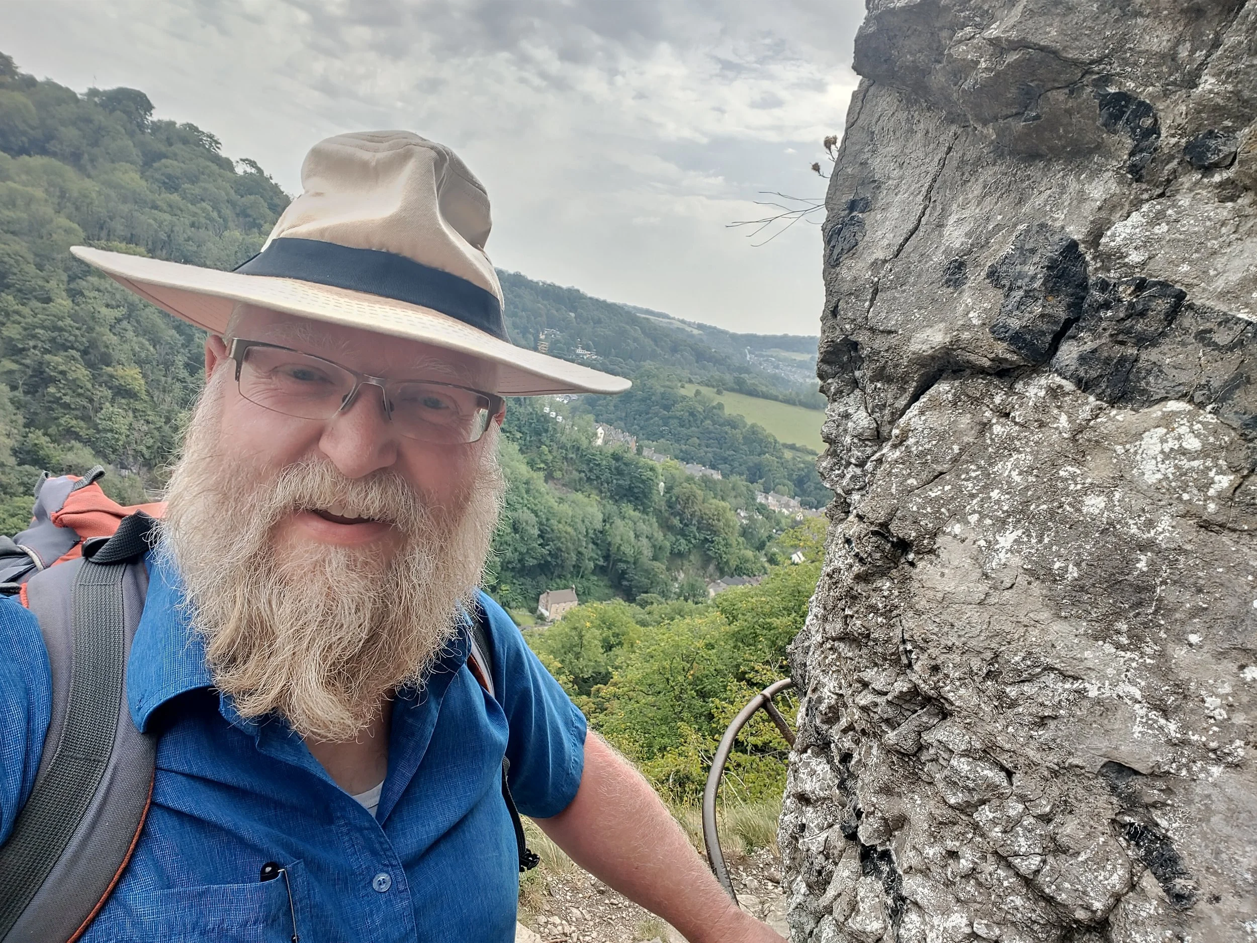 A man with a beard wearing glasses, a wide-brimmed hat, a blue shirt, and a backpack, smiling and standing next to a large rock formation outdoors with a green, hilly landscape in the background.