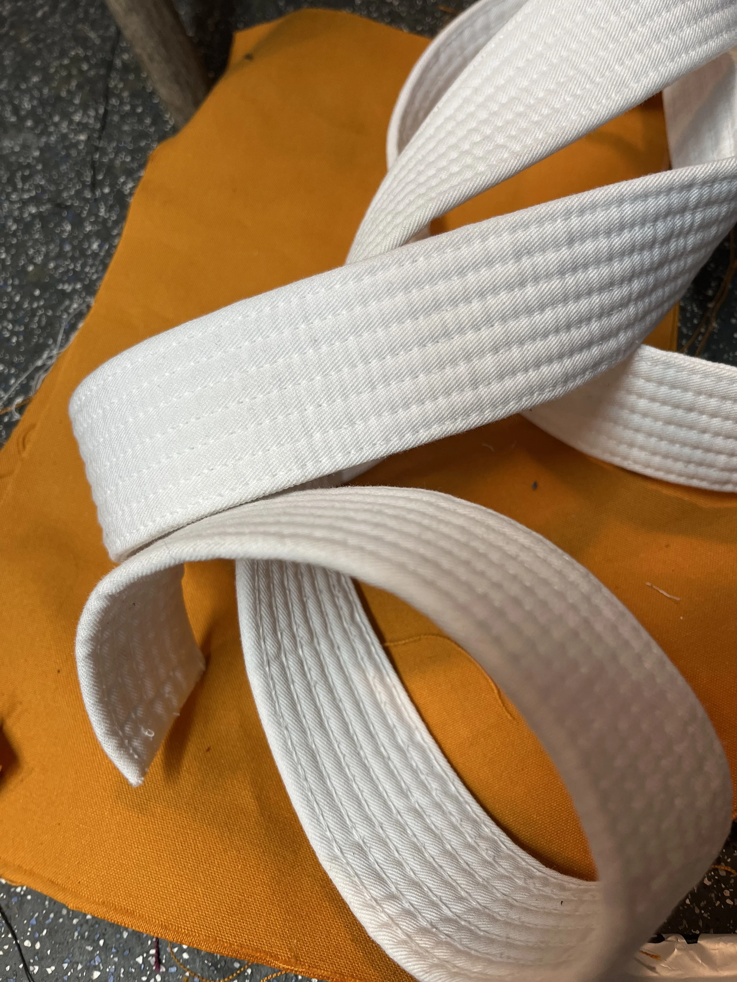 A close-up shot of white martial arts belts placed on an orange mat with a speckled black surface nearby.