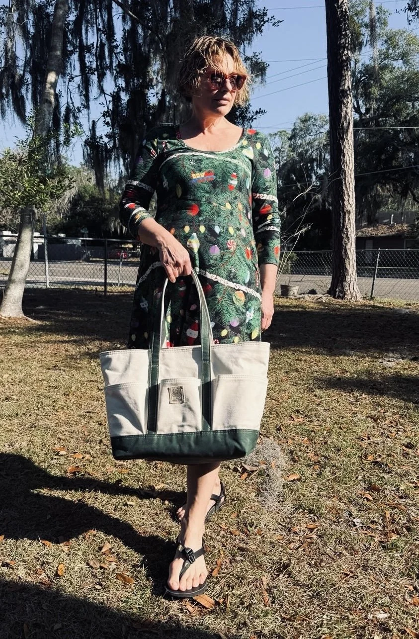 A woman standing outdoors on grass holding a large beige and green tote bag. She is wearing a colorful dress with a Christmas-themed pattern, sunglasses, and flip-flops. There are trees, a fence, and a street in the background with shadows cast on th