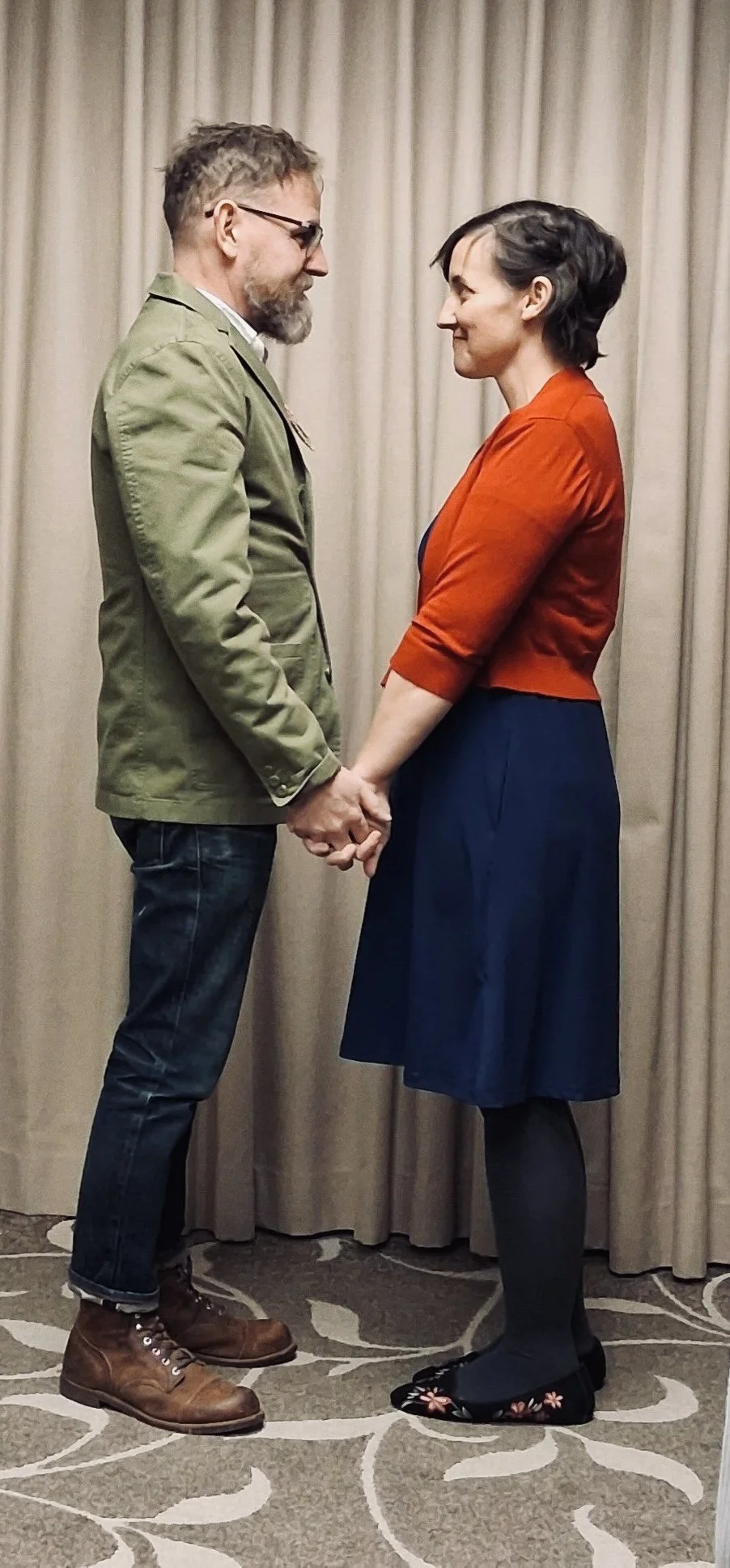A man and woman holding hands and gazing at each other indoors, standing on a patterned carpet with beige curtains in the background.