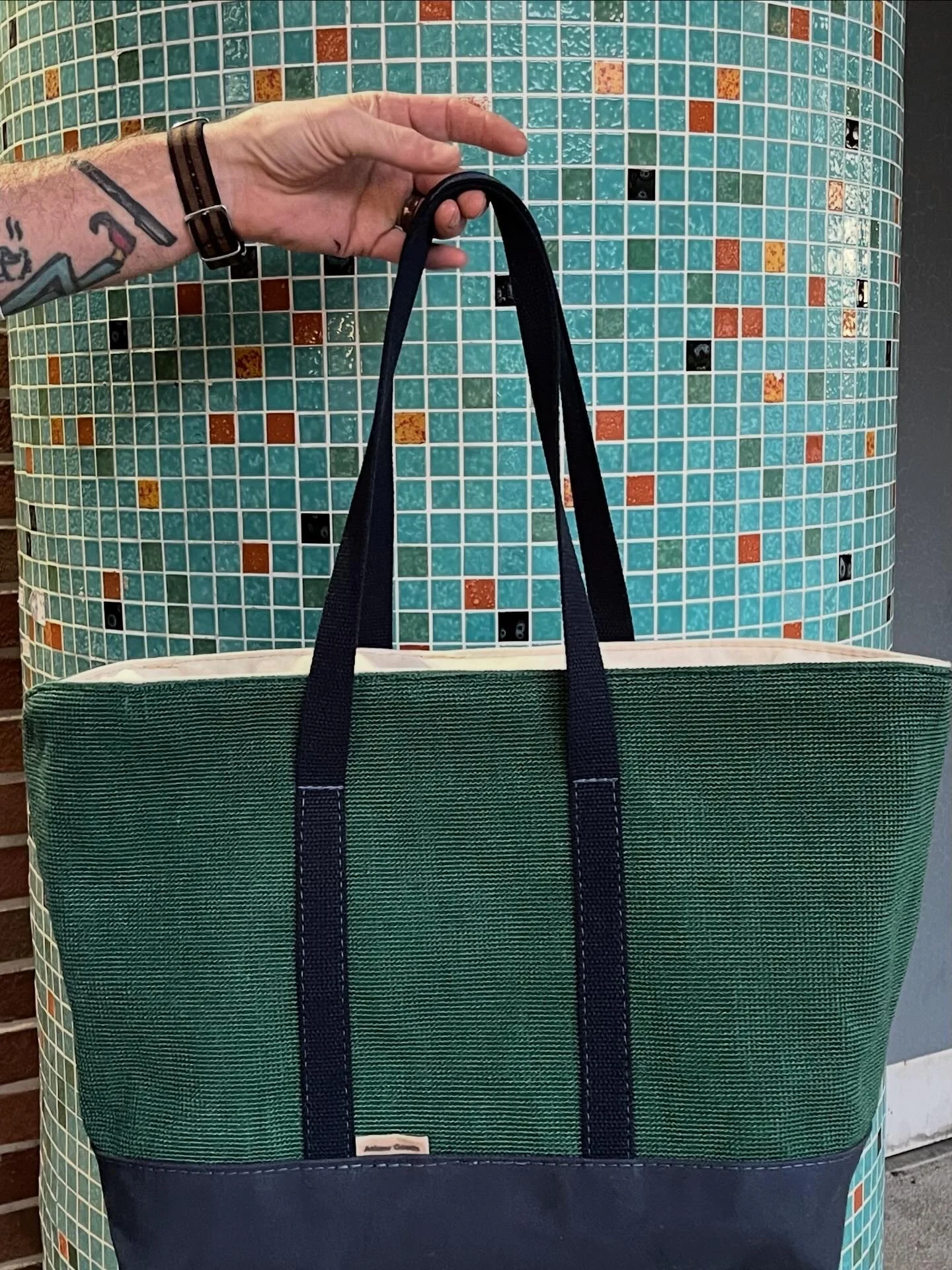 Person holding a large tote bag with green, black, and cream colors, against a tiled mosaic surface with small square tiles in shades of blue, orange, and black.