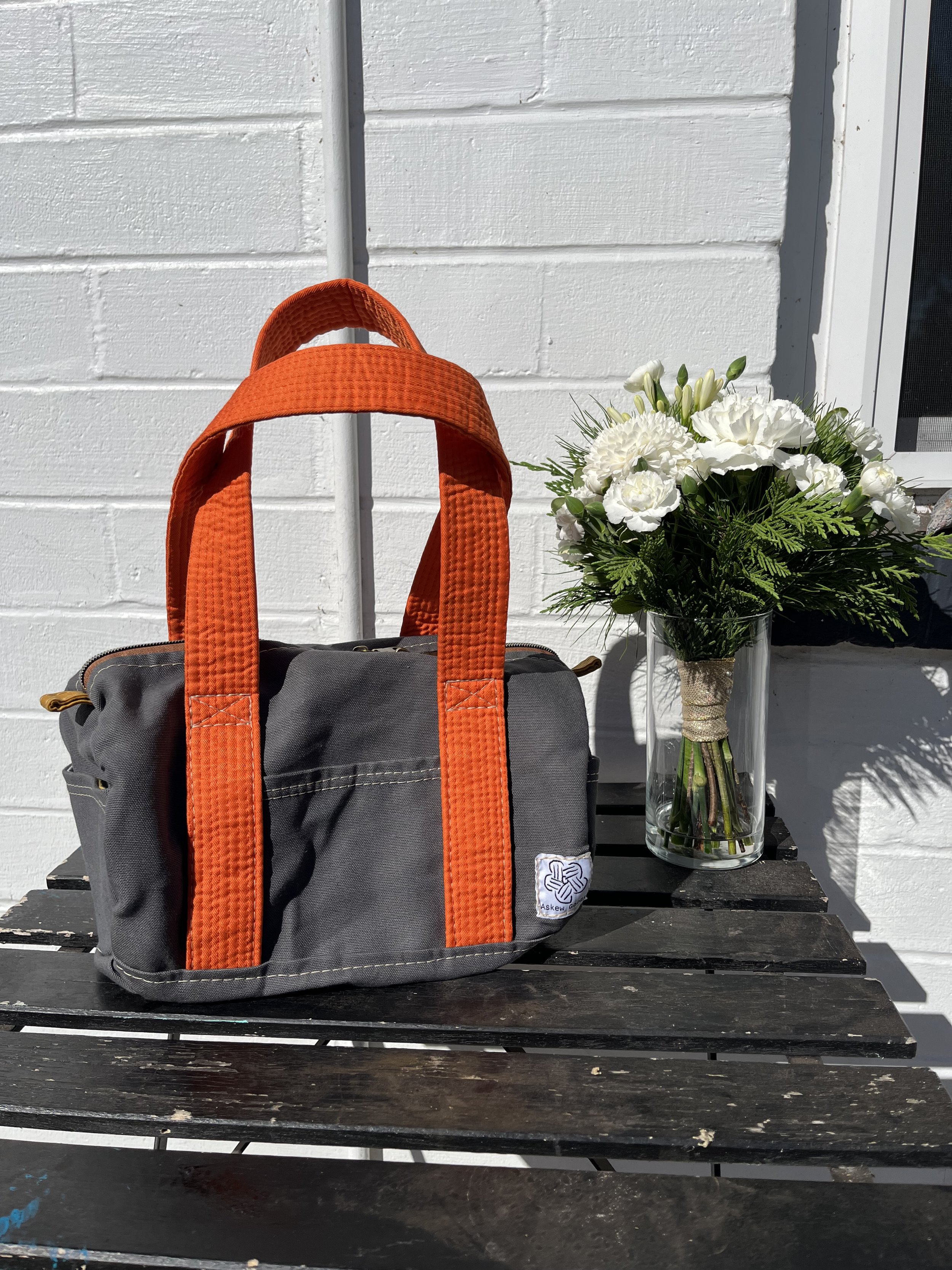 A black bag with orange handles sitting on a black wooden table, next to a glass vase filled with white flowers, outside against a white brick wall.