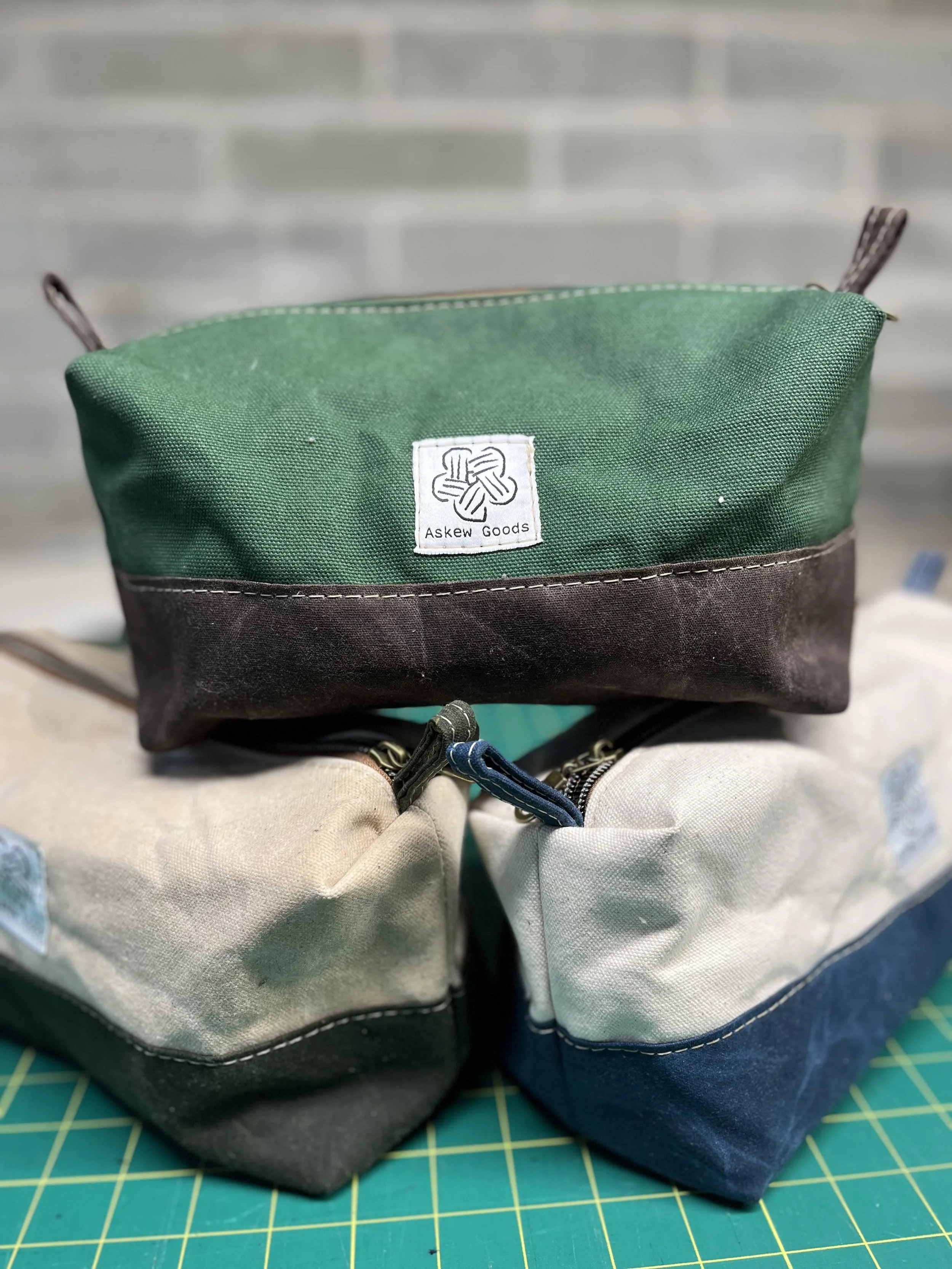 A multicolored fabric bag with a green upper section, dark brown middle section, and gray lower section, featuring a white square logo with the words "Askew Goods" and a knot design, placed on a cutting mat with a grid pattern.