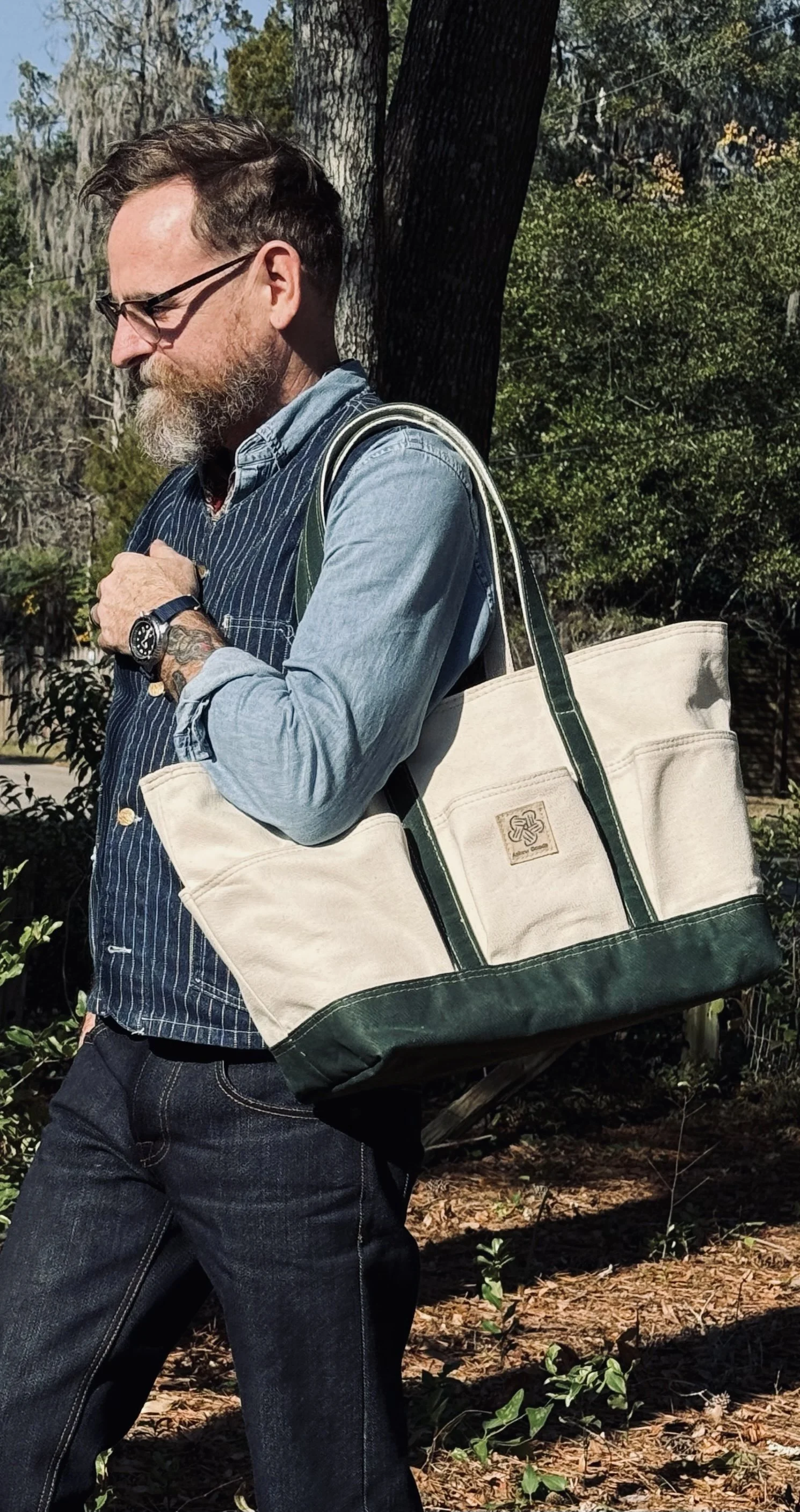 A man with glasses and a beard wearing a blue pinstripe shirt, carrying a large beige and green tote bag, walking outdoors near trees with sunlight.