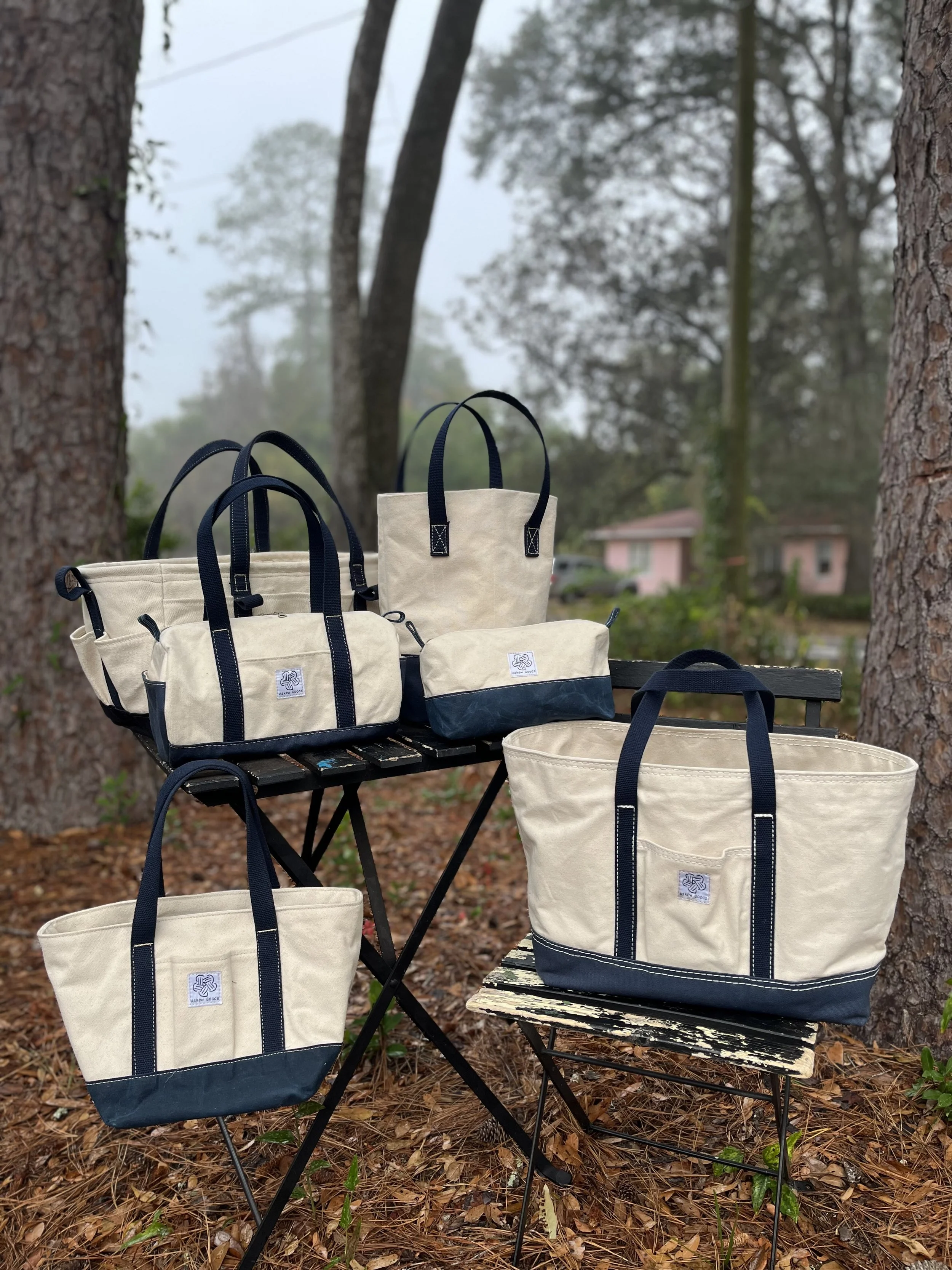 Several cream-colored canvas tote bags with navy handles and accents displayed outdoors on a black metal table and chair, surrounded by trees and fallen leaves.